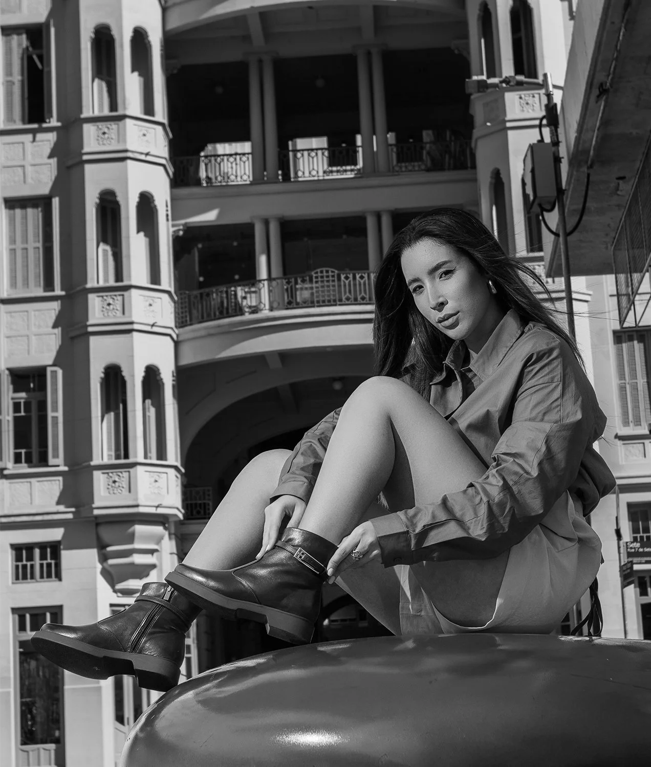 Retrato de uma mulher com cabelo solto, vestindo jaqueta e botas de couro, sentada em uma estrutura metálica em frente a um prédio antigo, em uma cidade, em uma foto em preto e branco.