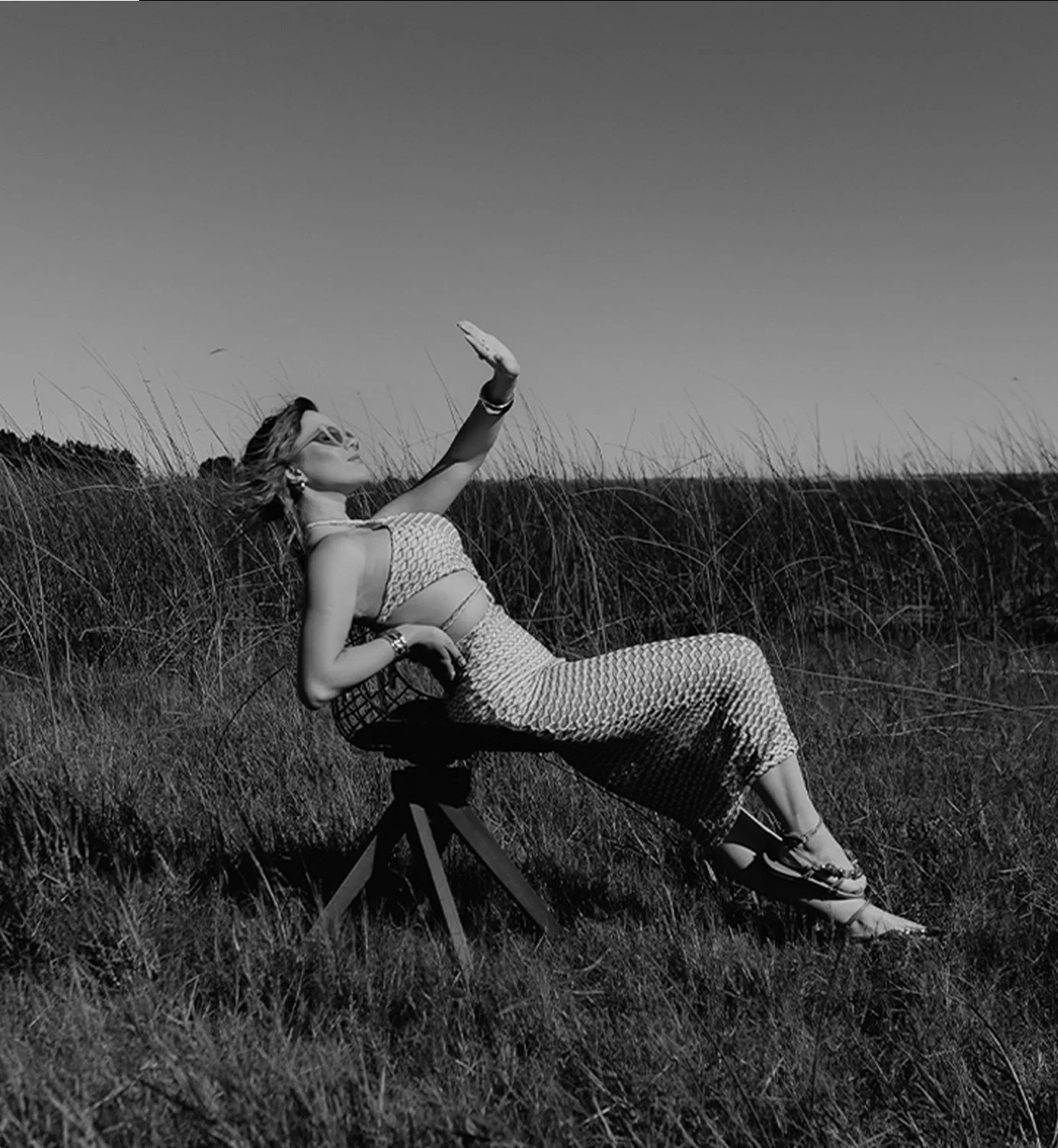 Mulher relaxando em um campo, sentada em uma cadeira, usando vestido e óculos de sol, com o braço levantado, em um cenário ao ar livre.