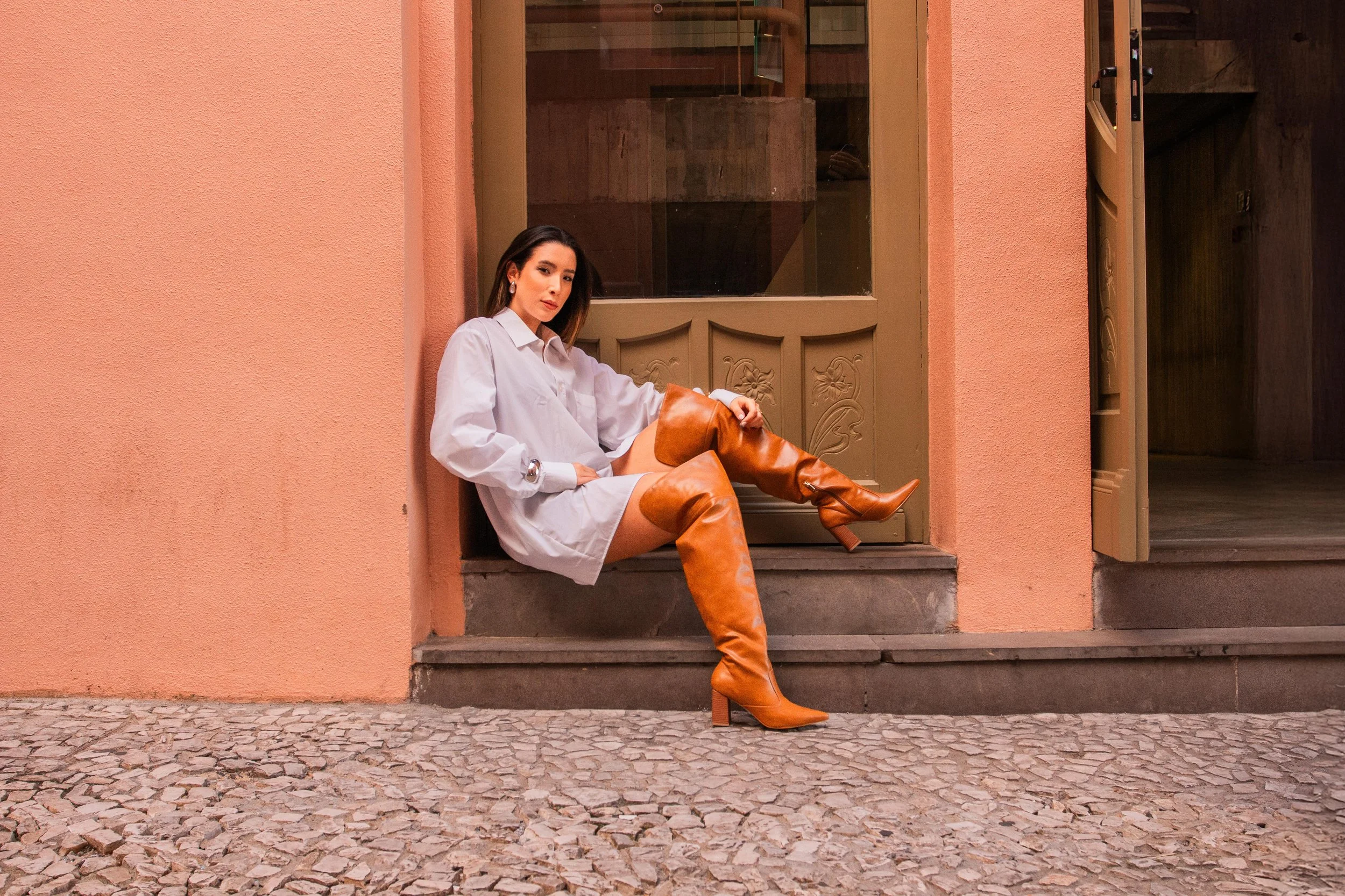 Mulher de cabelo castanho escuro usando uma blusa branca e botas altas de couro marrom, sentada na entrada de uma porta de madeira com moldura decorativa, ao lado de uma parede de cor pêssego.