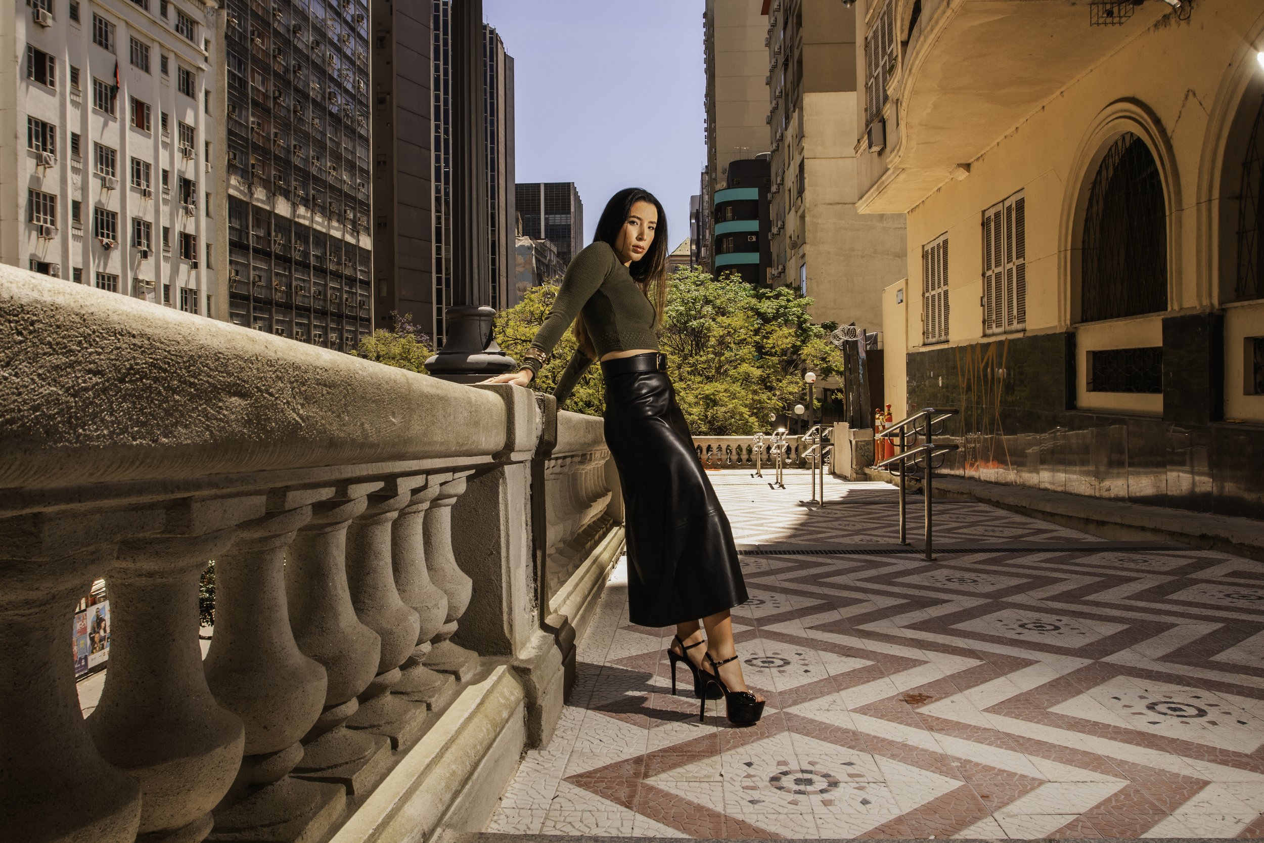 Mulher usando saia longa de couro preto, blusa de manga comprida verde, e salto alto, posando em um passeio urbano entre edifícios, com árvores ao fundo.