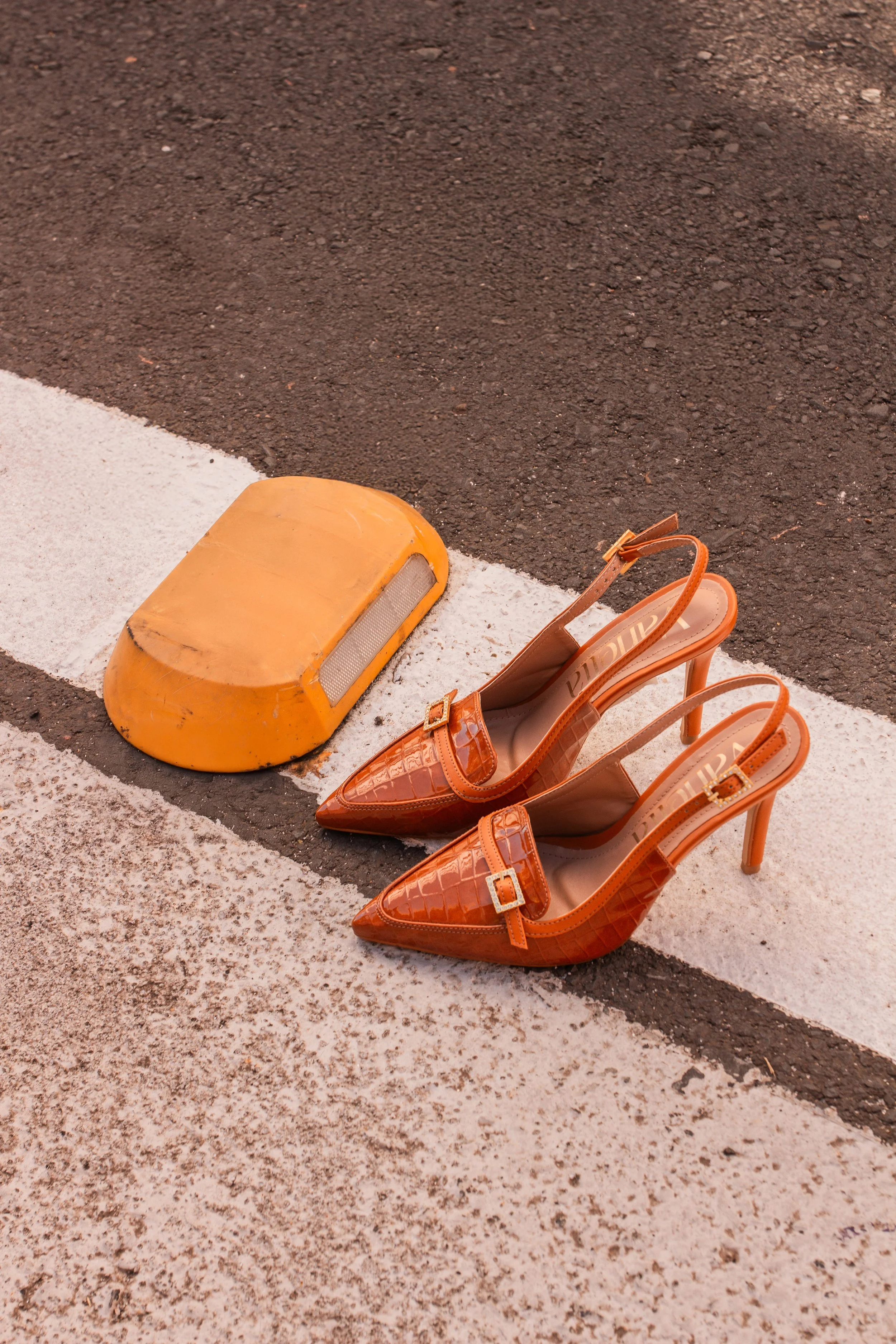 Sapatos de salto alto laranja com bico pontudo, detalhes de couro texturizado e fivelas, um par de sapatos posicionado no chão de asfalto, ao lado de um equipamento de tráfego amarelo.