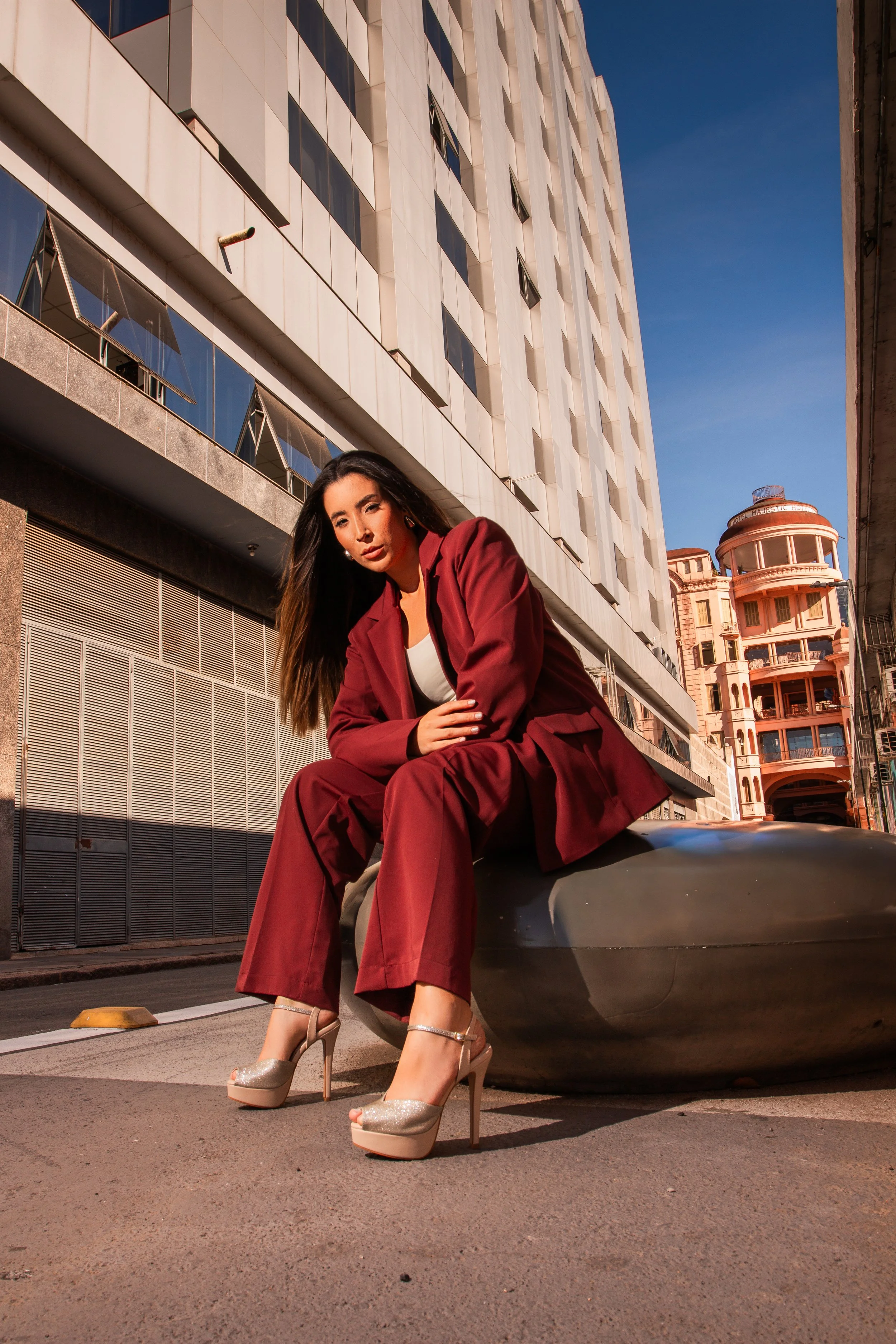 Mulher jovem com cabelo castanho escuro, vestindo terno vermelho e salto alto dourado, sentada em uma estrutura de arte urbana em uma rua de cidade.