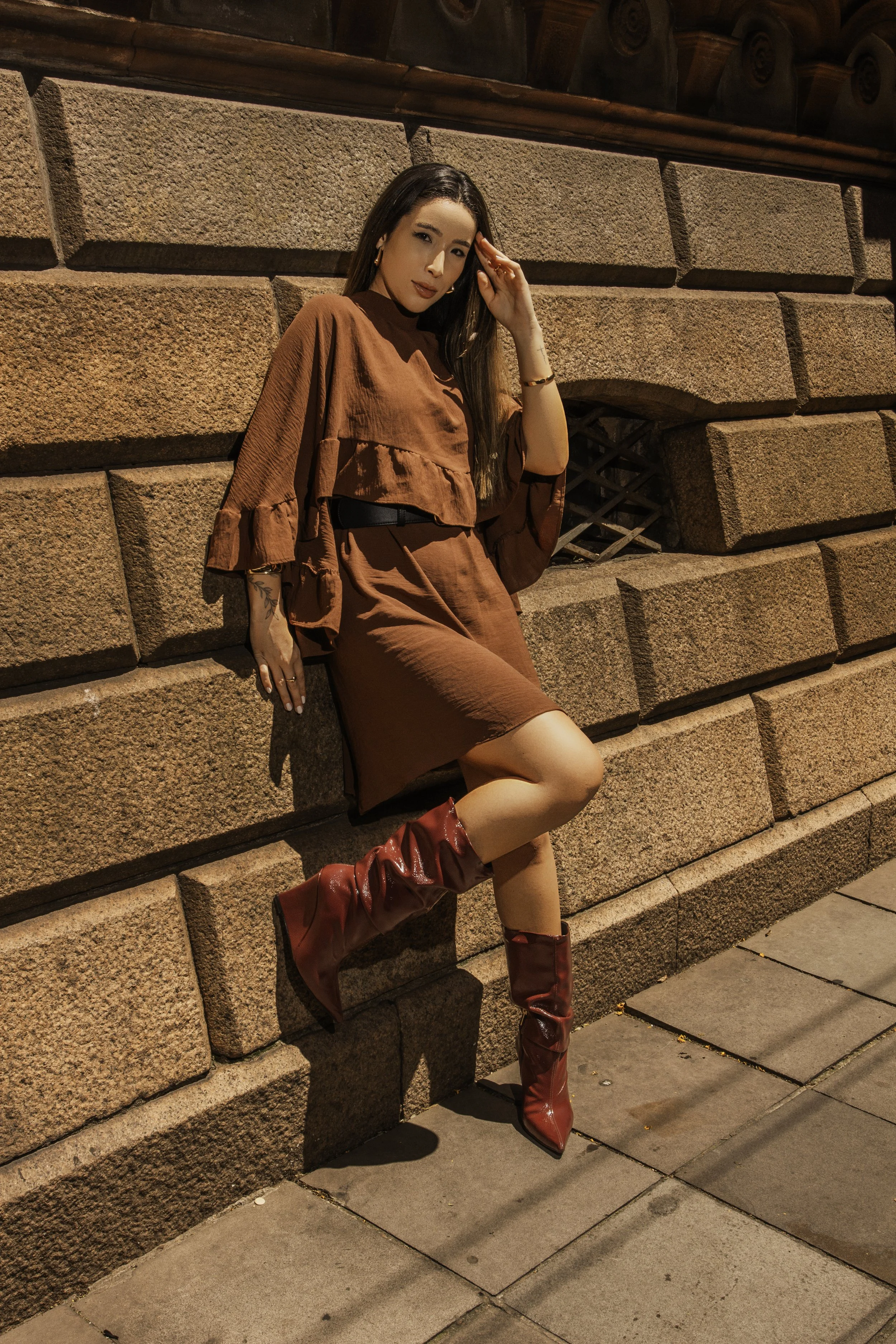 mulher posando contra parede de pedra, usando roupa marrom e botas vermelhas