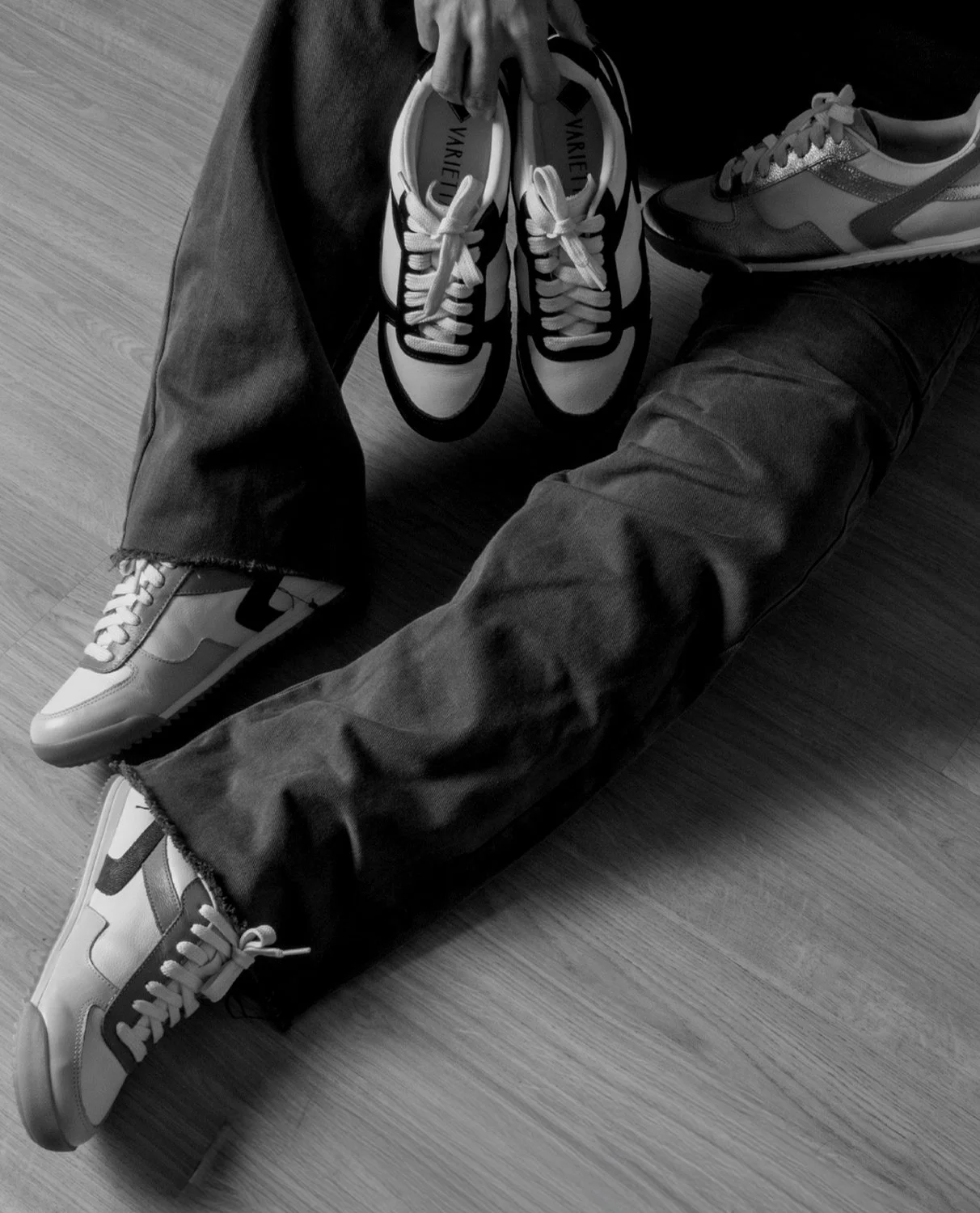 Pessoa com jeans escuros sentado no chão, segurando um par de tênis esportivos com detalhes pretos e brancos, enquanto outra pessoa está com uma tênis com cores variadas.