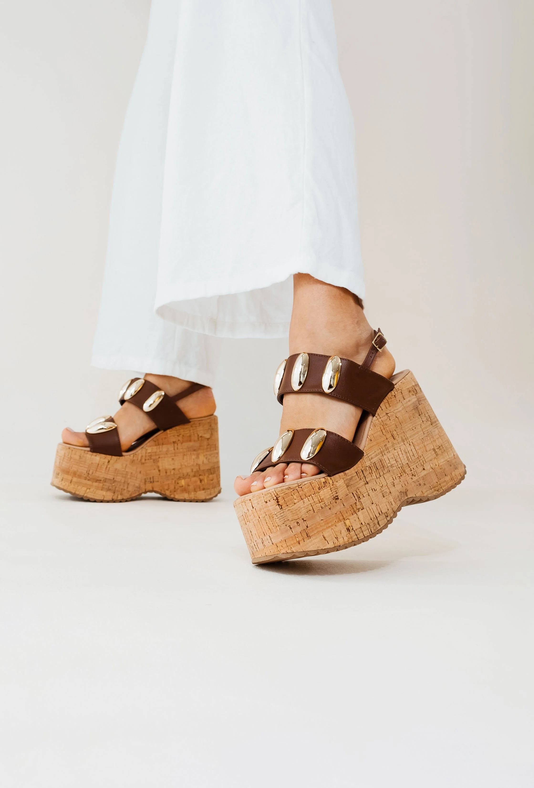 Sandálias plataforma de cortiça com tiras de couro marrom e detalhes dourados, usadas por uma pessoa com vestido branco.
