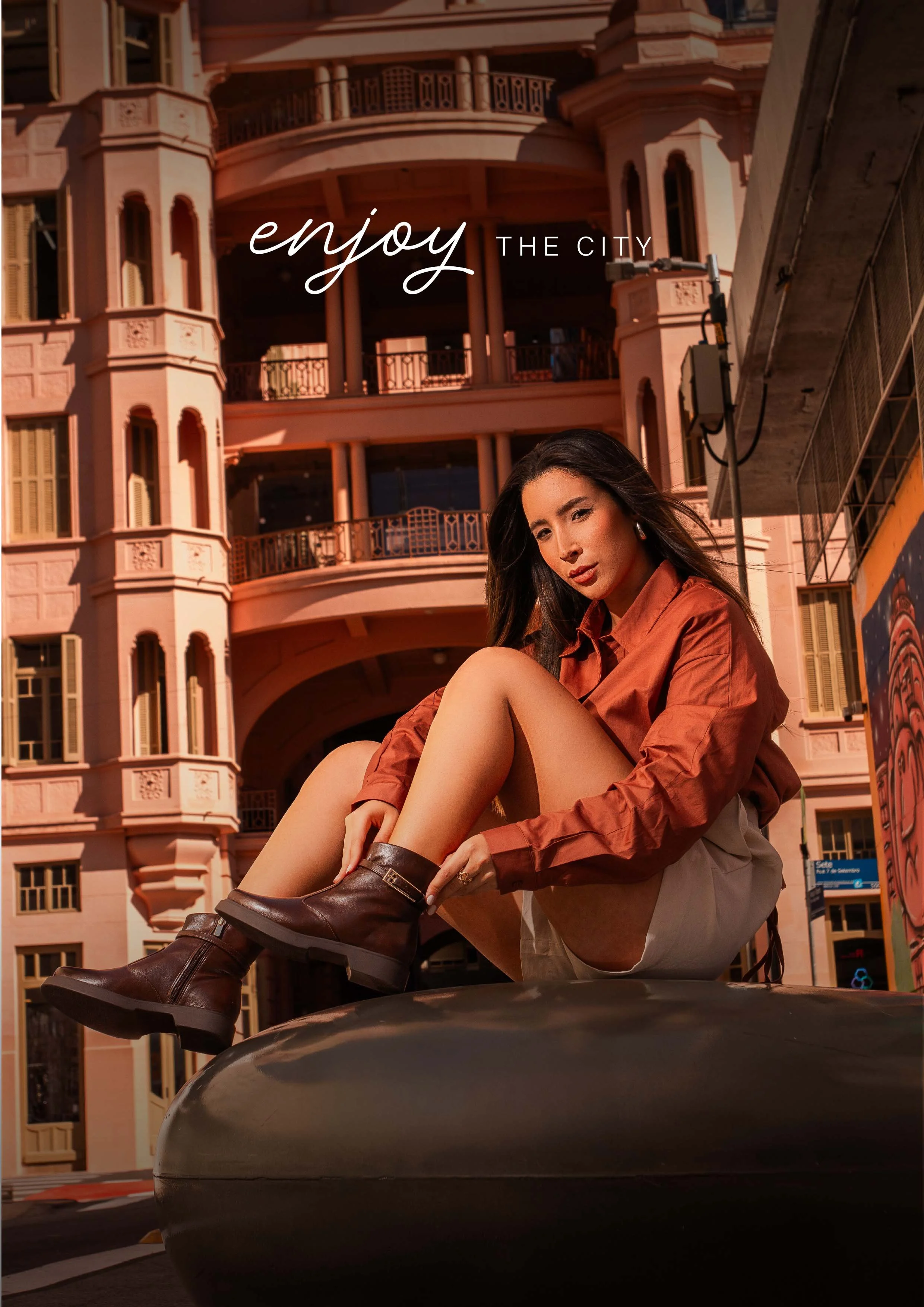 Mulher jovem sentada sobre uma estrutura em uma rua urbana, com prédio de arquitetura clássica ao fundo, usando jaqueta marrom e botas, ao pôr do sol.