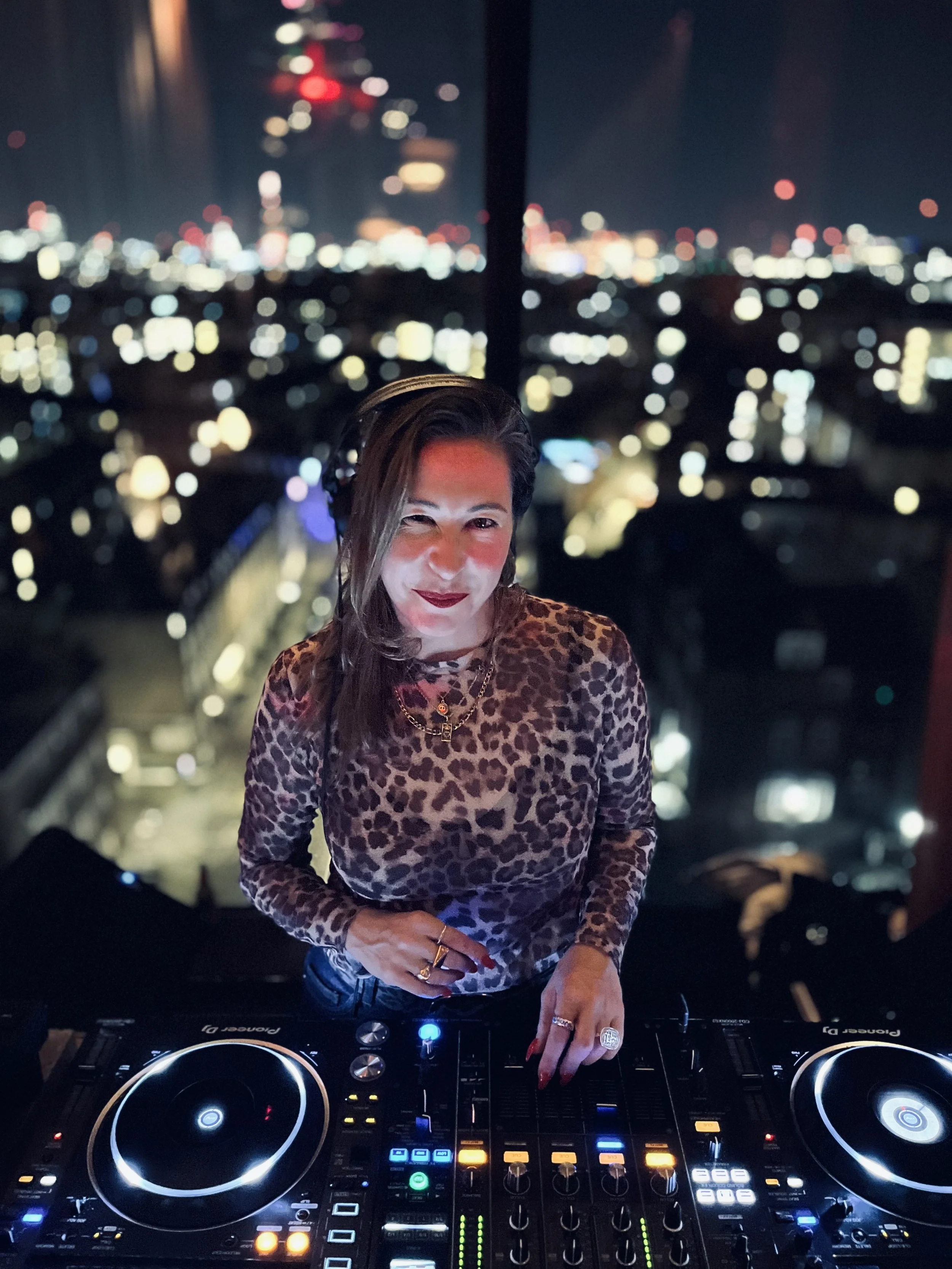A woman with leopard print clothing and jewelry is DJing at a club, with a city skyline lit up at night in the background.