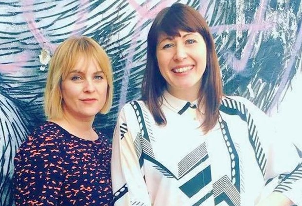 Two women standing in front of a tropical leaf mural, smiling and posing for the photo.
