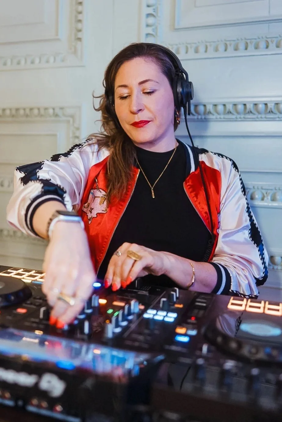 A woman DJ wearing headphones and a red and white jacket, working with DJ equipment in a room with ornate white walls.