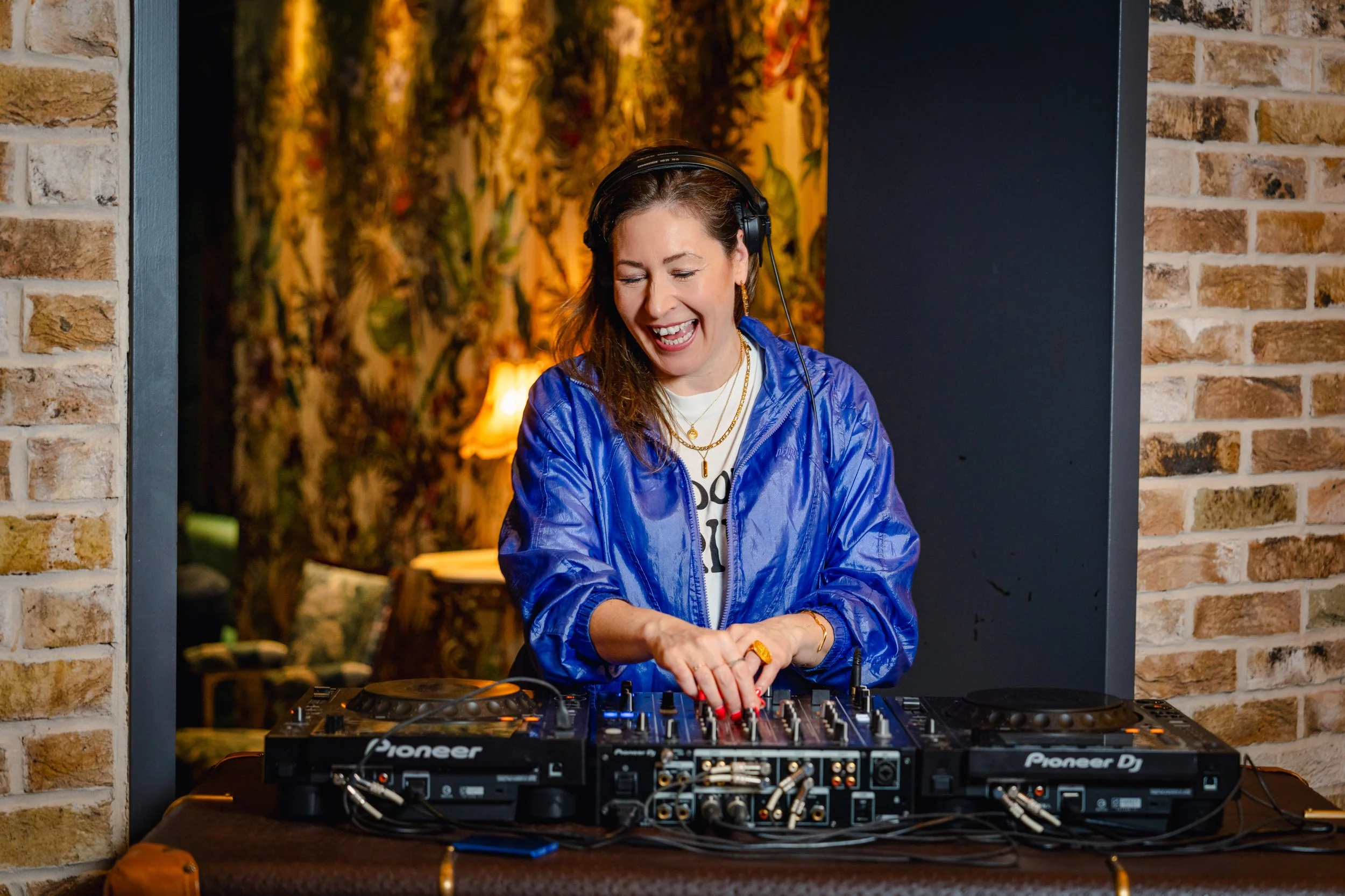 A woman with brown hair, wearing a blue jacket, and headphones, DJing with a Pioneer DJ mixer in a warmly lit room with brick walls and floral decorations in the background.