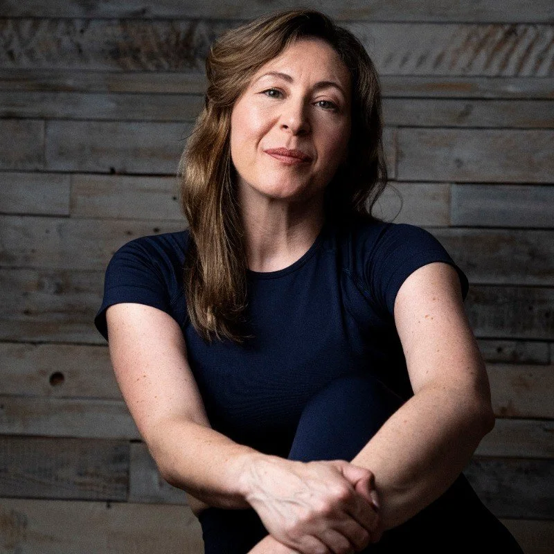 A woman with wavy brown hair wearing a navy blue t-shirt, sitting casually against a wooden panel wall, with her arms crossed over her legs, looking at the camera with a slight smile.