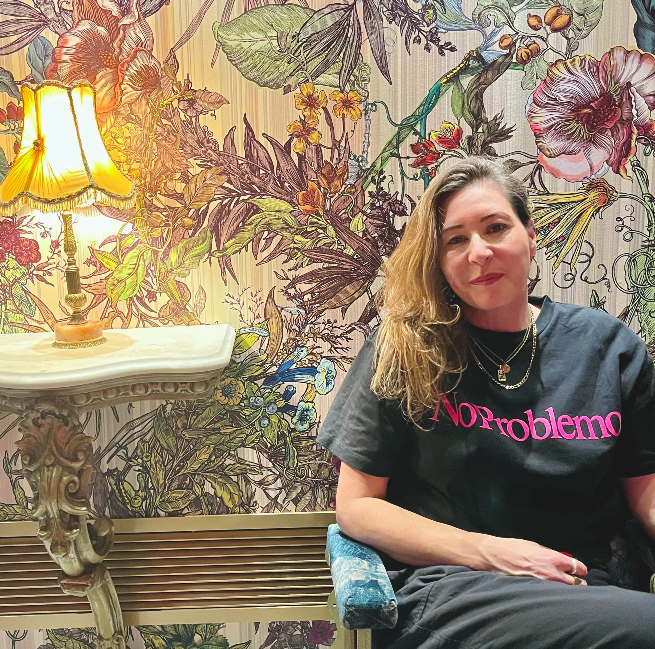 A woman with shoulder-length wavy hair and earrings, sitting in front of a large floral mural, wearing a black T-shirt with pink text and silver jewelry, with a table lamp on her left.