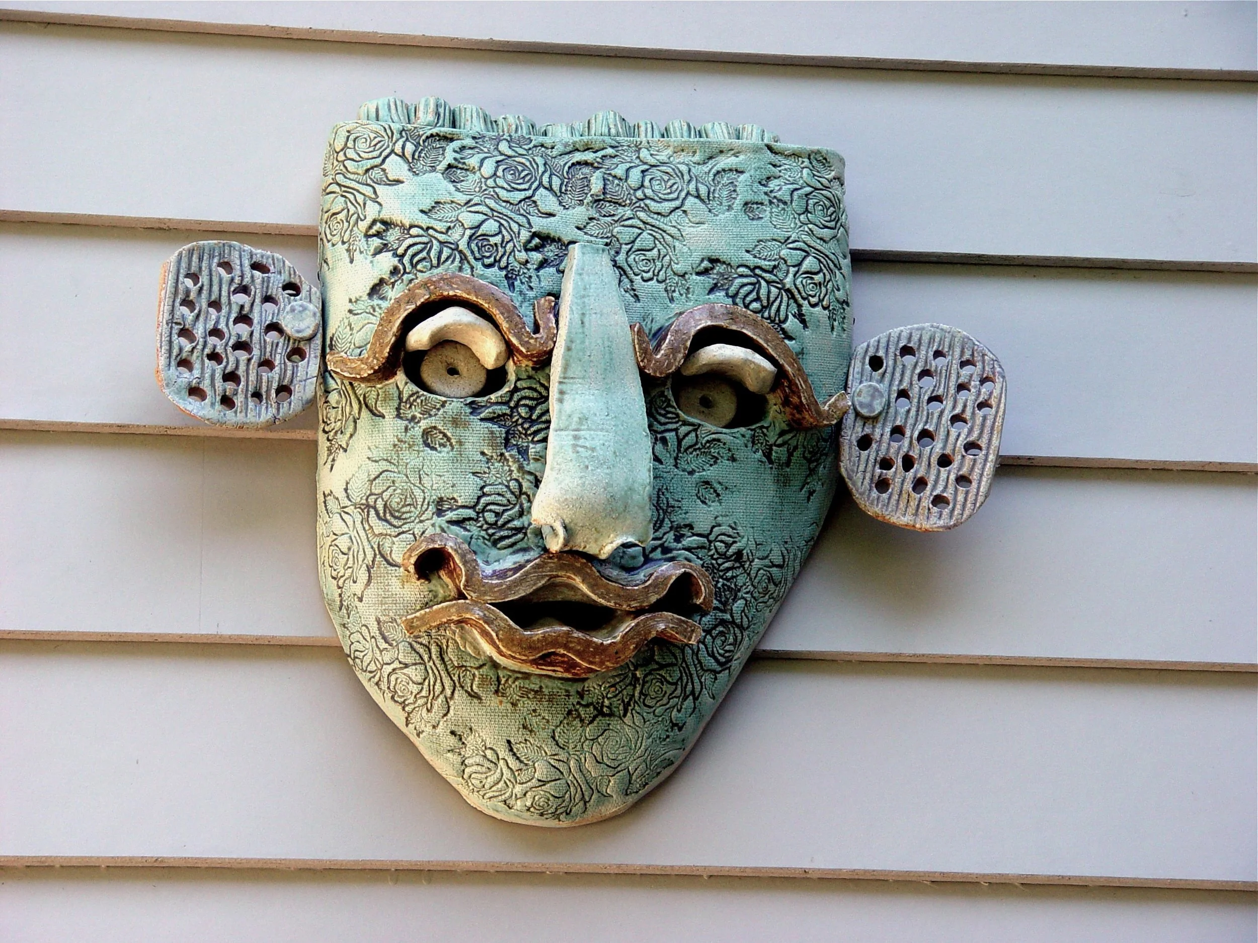 Decorative ceramic face mask with components resembling elephant ears, mustache, and lips, mounted on a wall with horizontal siding.