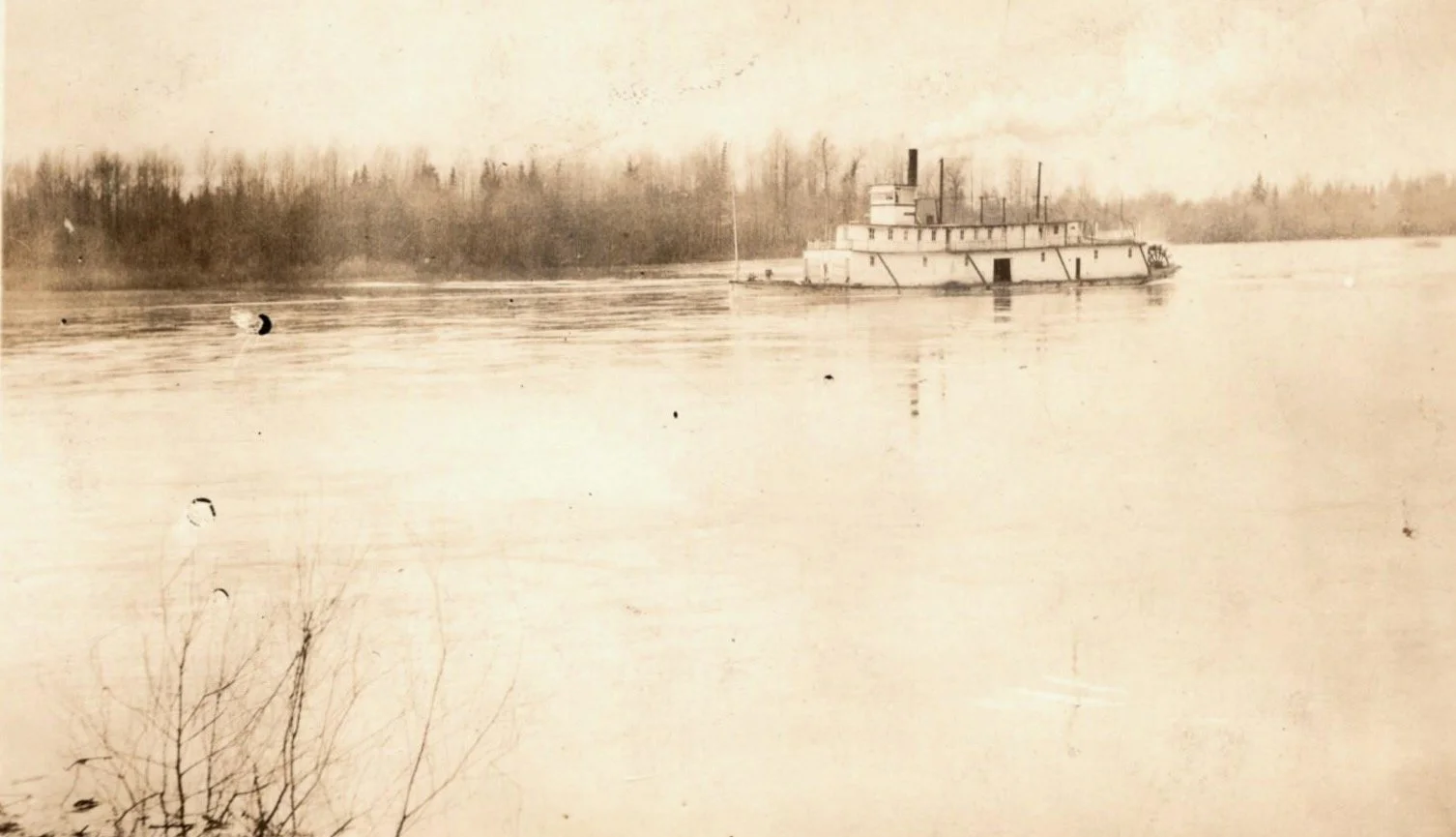 An old sepia-toned photograph of a river with a large steamboat in the distance and a forested shoreline in the background.