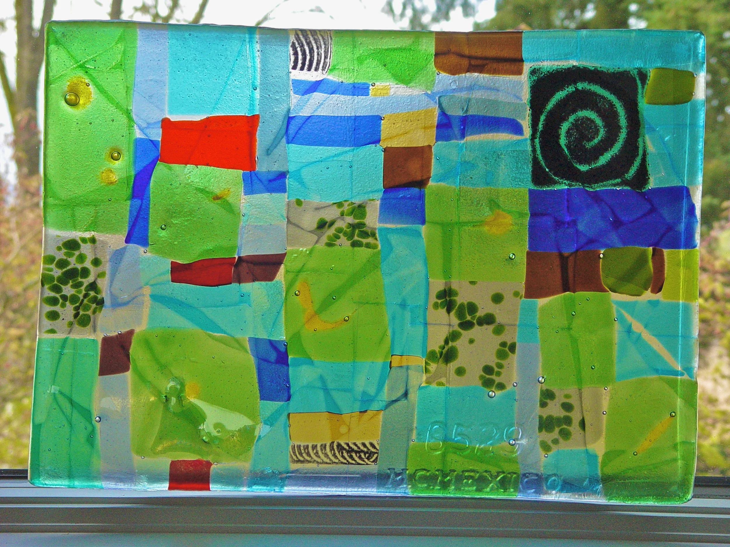 A colorful stained glass window with various abstract shapes and colors, including green, blue, red, yellow, and black spiral design, with sunlight shining through.