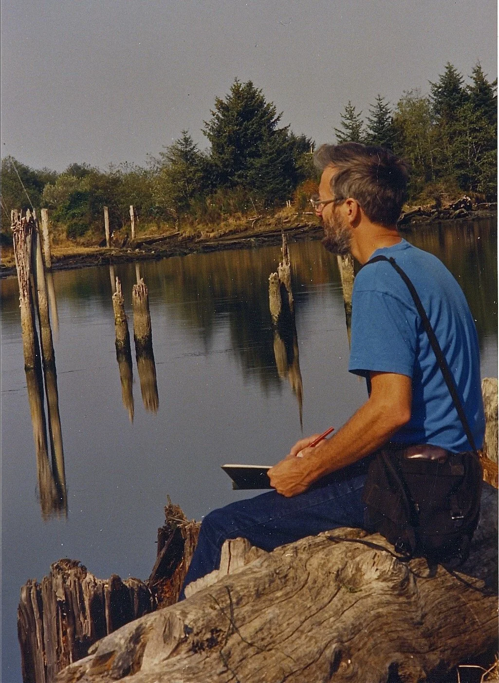 A man with glasses and a beard, dressed in a blue T-shirt, sitting on a large piece of driftwood by a calm river. He is holding a notebook and writing, with weathered wooden posts sticking out of the water nearby. The background features a forest wit