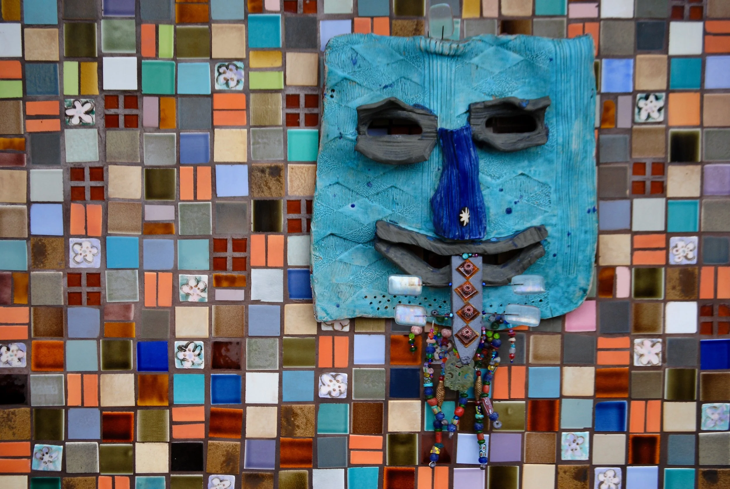 A mixed-media art piece featuring a face made from painted wood, positioned against a colorful mosaic tile background. The face has exaggerated features, including a prominent blue nose, black eye frames, and a wide smiling mouth with necklaces hangi