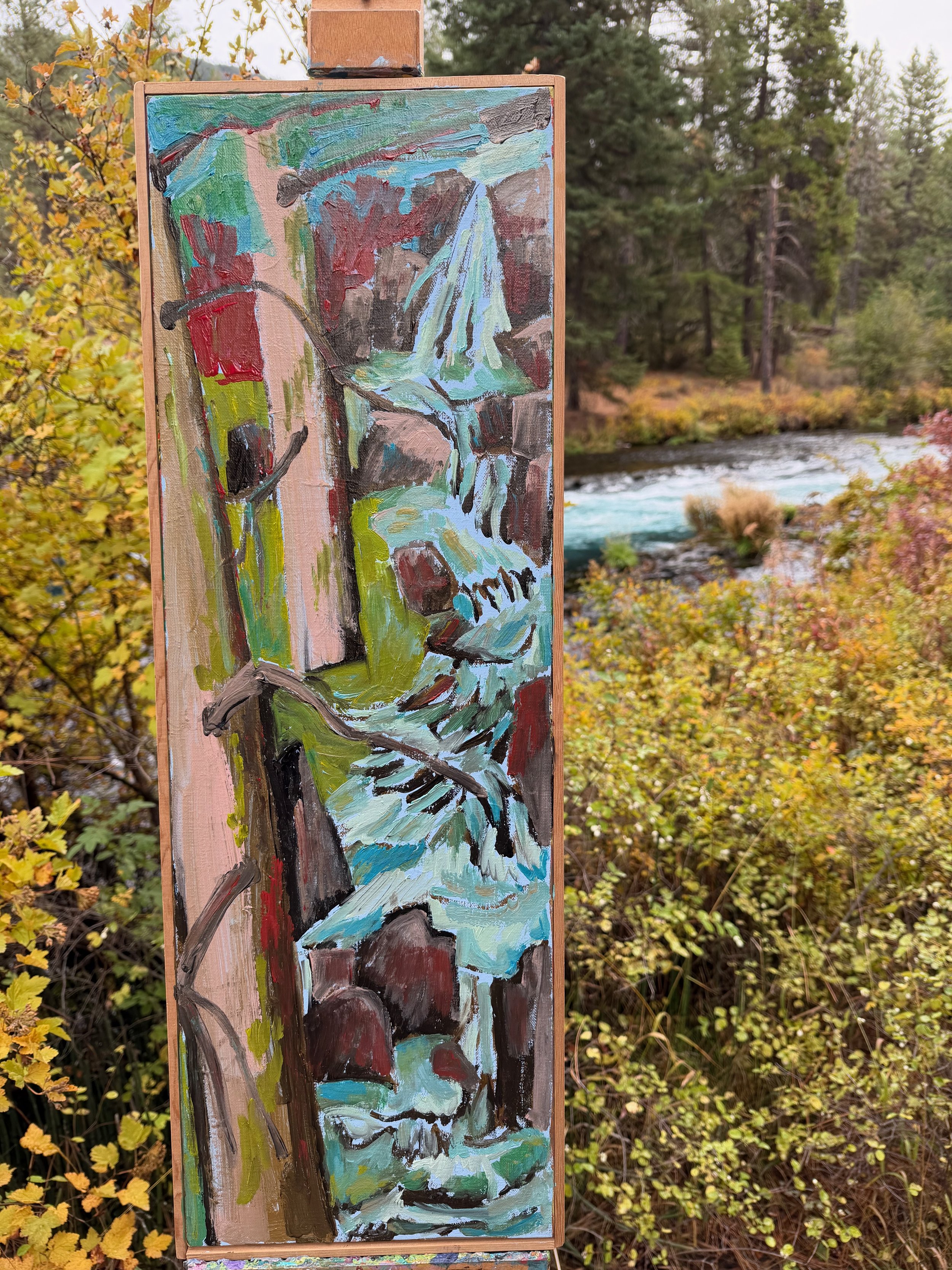 A colorful abstract painting of a waterfall on a vertical canvas, set outdoors with a river and trees in the background.