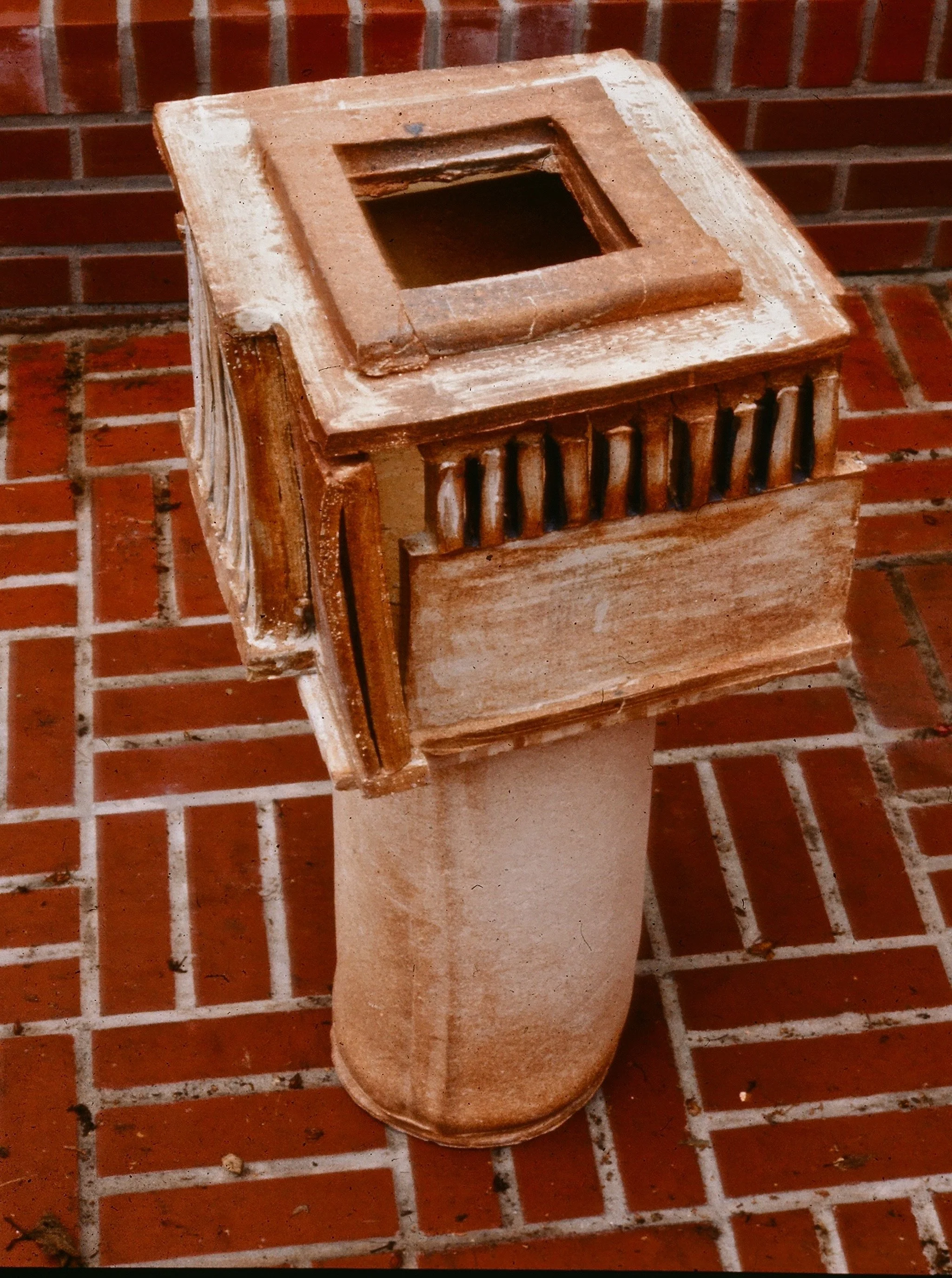 A terracotta chimney with a square opening on top, sitting on a brick patio.