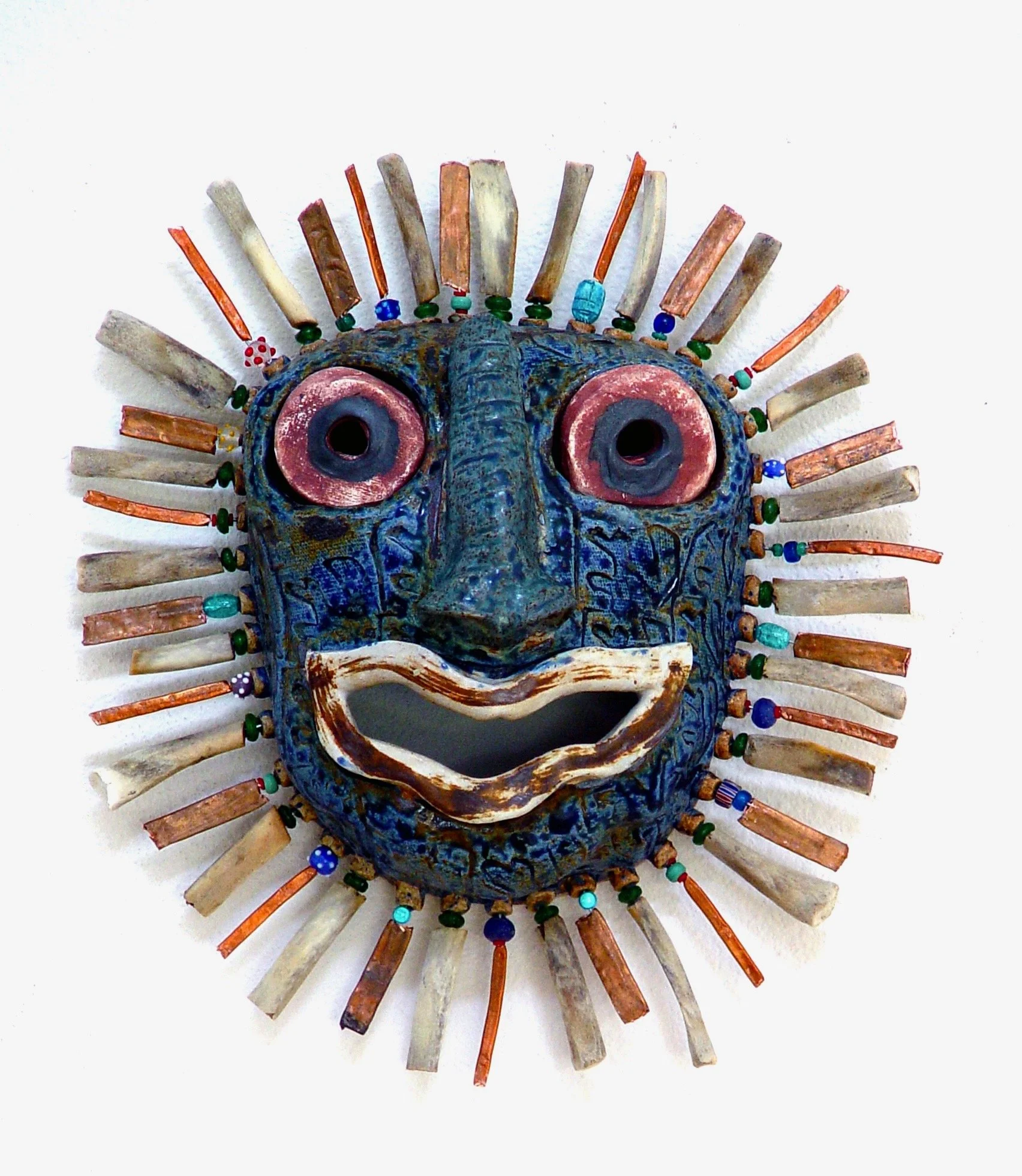 A colorful mask with a face, large eyes, a wide open mouth, surrounded by beaded and wooden strips that resemble a sunburst.