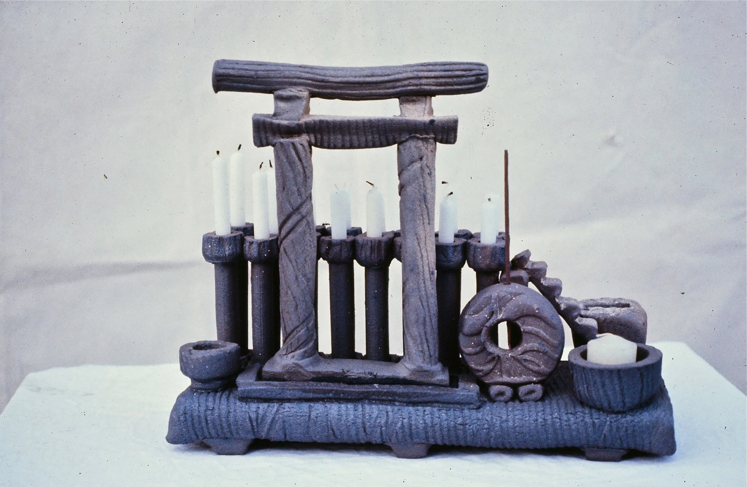 Wooden miniature Japanese torii gate with six white candles on black holders and a wooden wheel, set on a stone-textured base.