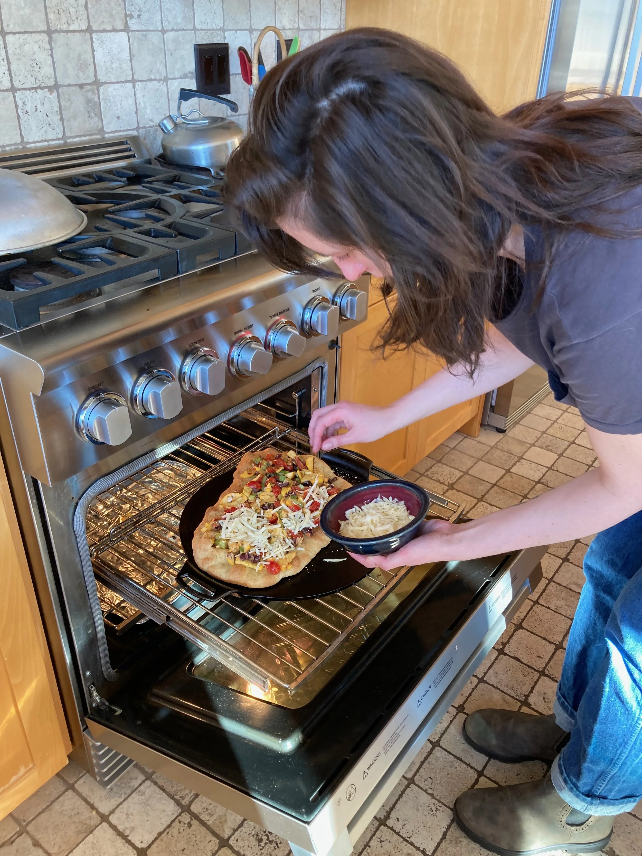 Person placing a freshly baked pizza into an oven, holding a bowl of shredded cheese.