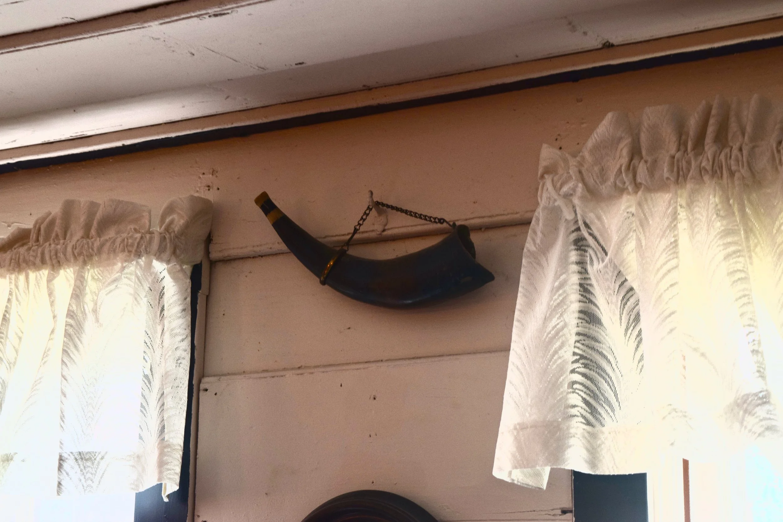 Horn-shaped decorative wall hanging with chain attachment between two windows with white floral curtains in a rustic interior.