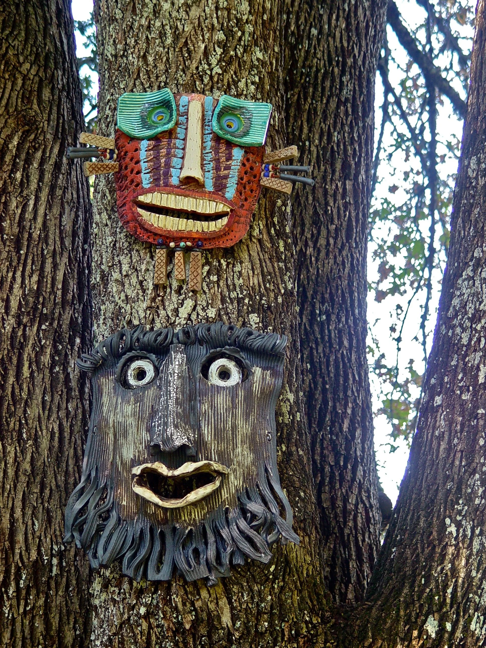 Two colorful, artistic masks mounted on a tree trunk, one above the other.