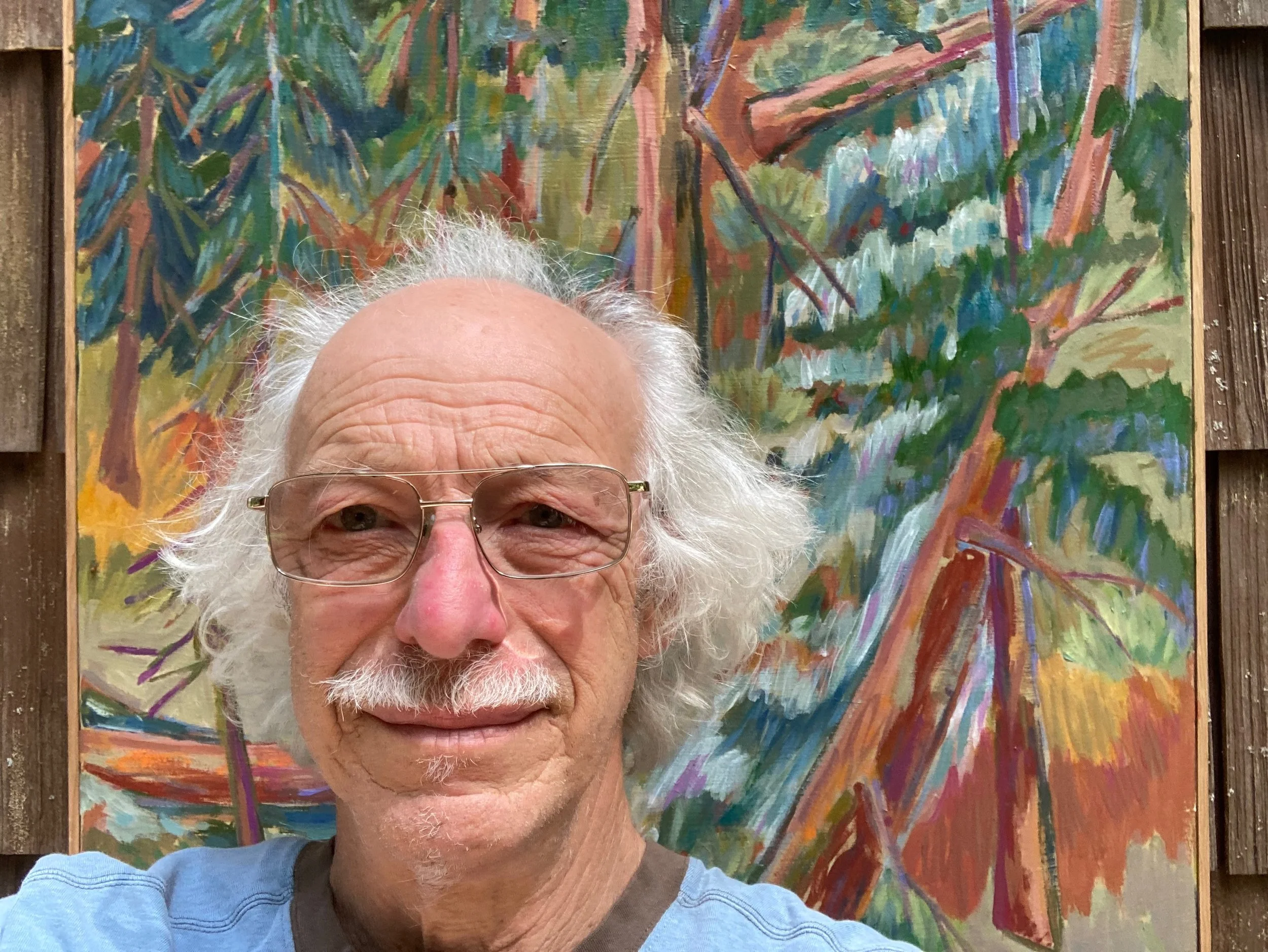 An older man with white curly hair, glasses, and a mustache taking a selfie in front of a colorful abstract painting with leafy patterns, surrounded by wooden panels.