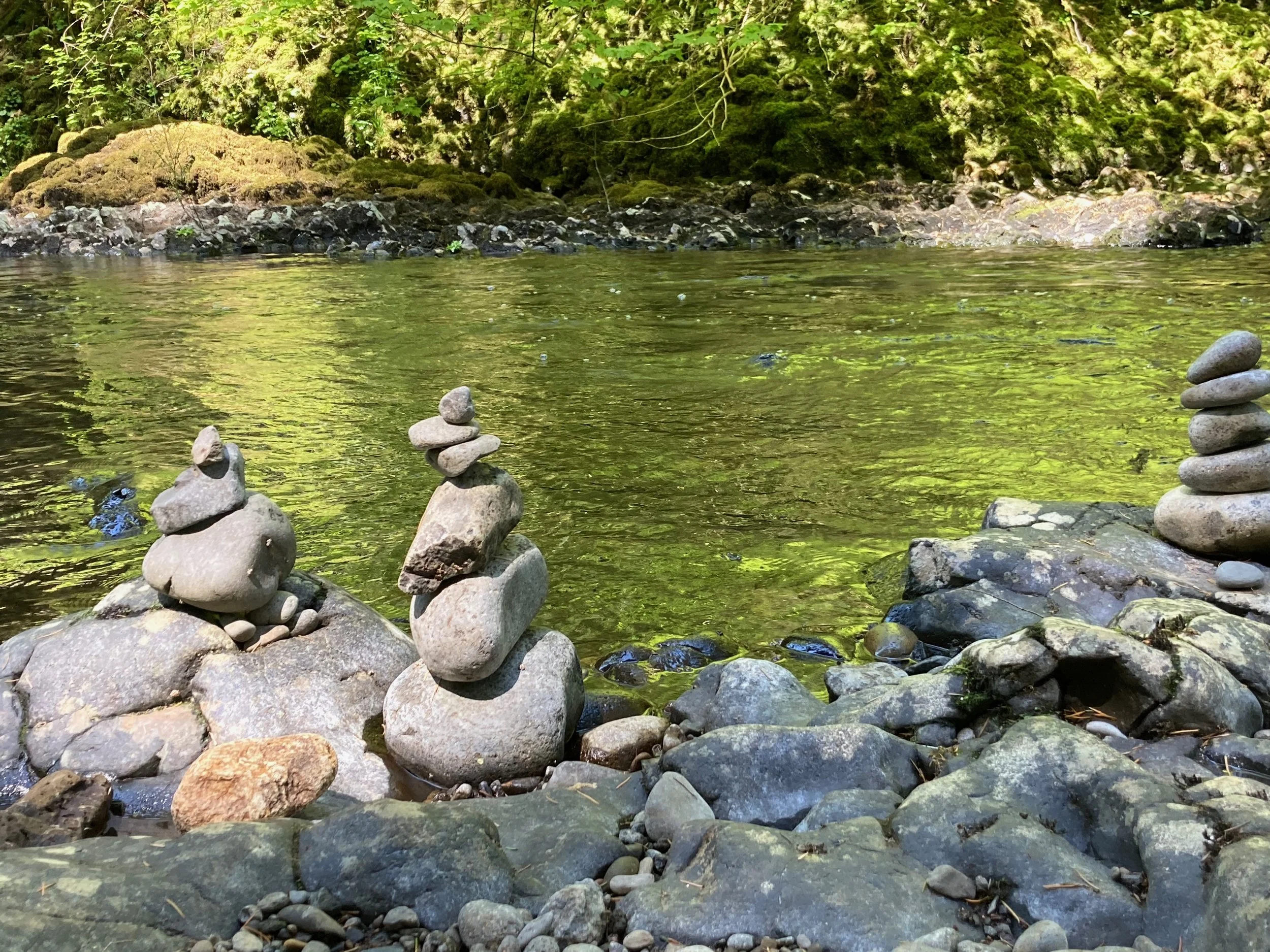 Stacks of rocks arranged on a rocky riverbank near flowing water with green foliage in the background.