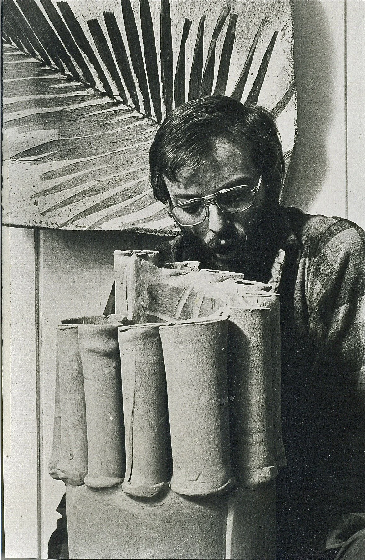 A person with glasses and a beard leans over a surface, looking at a large collection of cylindrical objects, possibly art or craft materials, with a leaf pattern artwork hanging on the wall behind them.