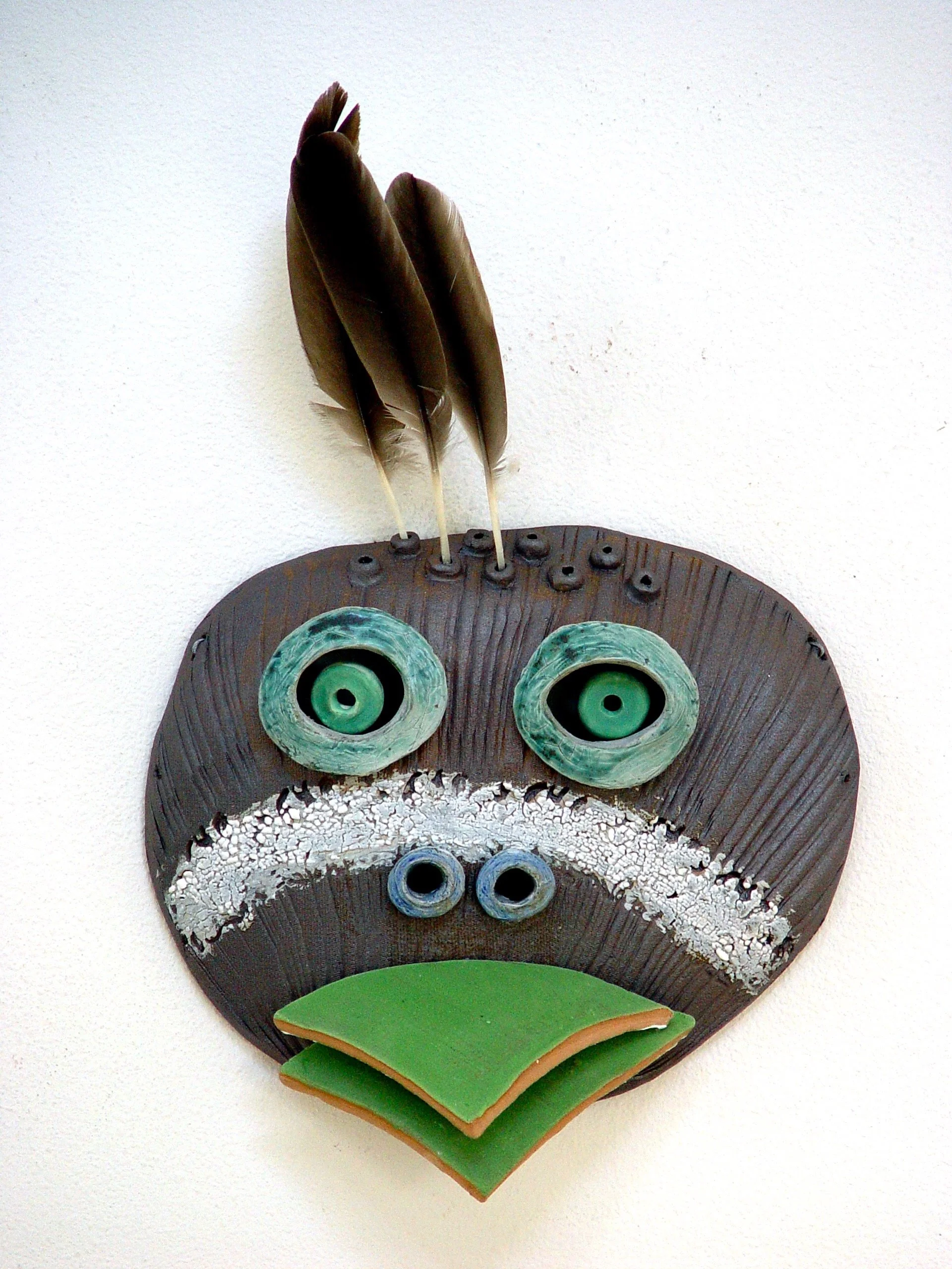A decorative mask resembling a frog face, with feather hair, large round eyes, a white textured mustache, and a green and tan mouth, mounted on a white wall.