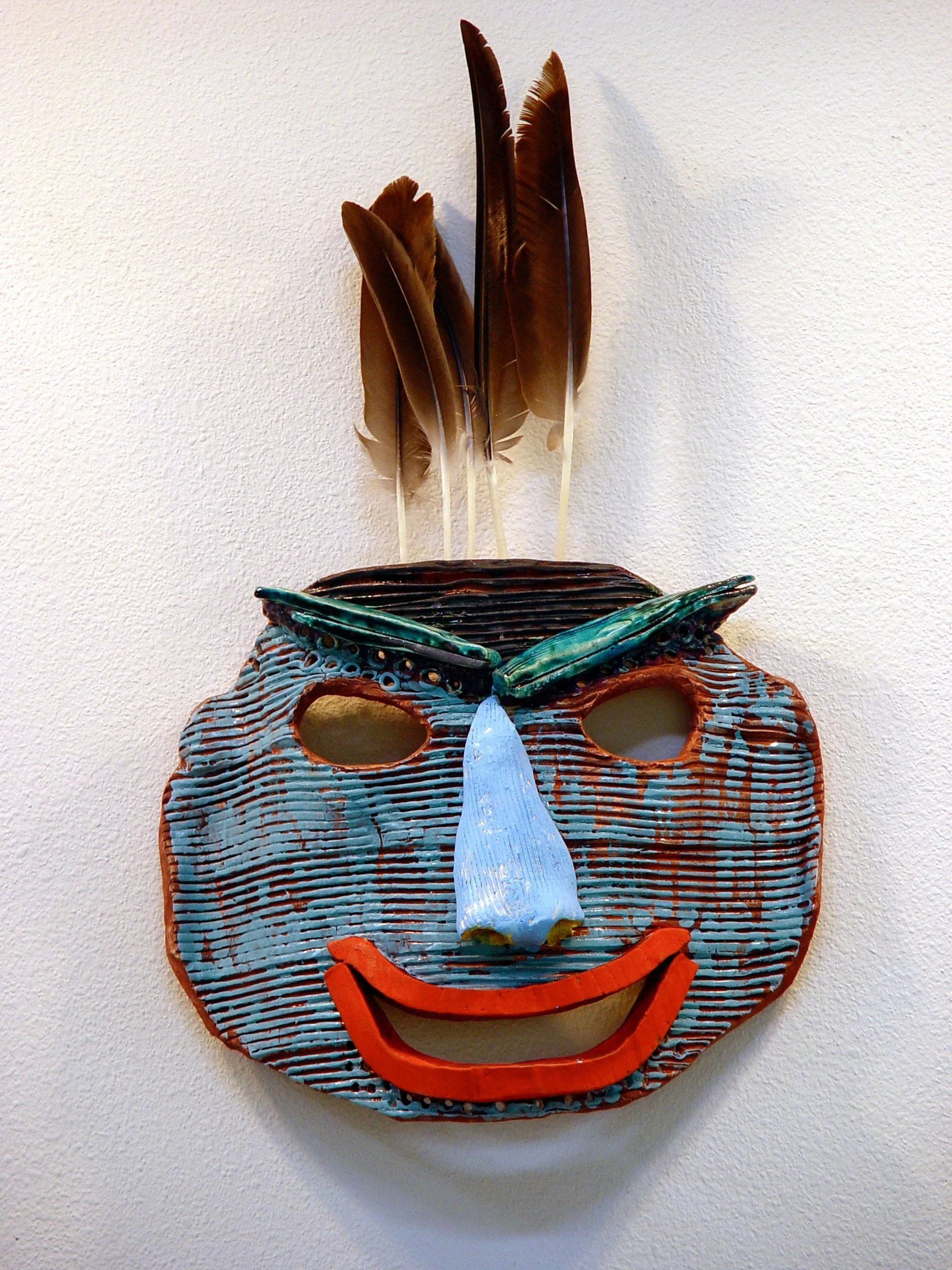 Decorative mask resembling a smiling face with feathers on top, made of colorful ceramic and wood.