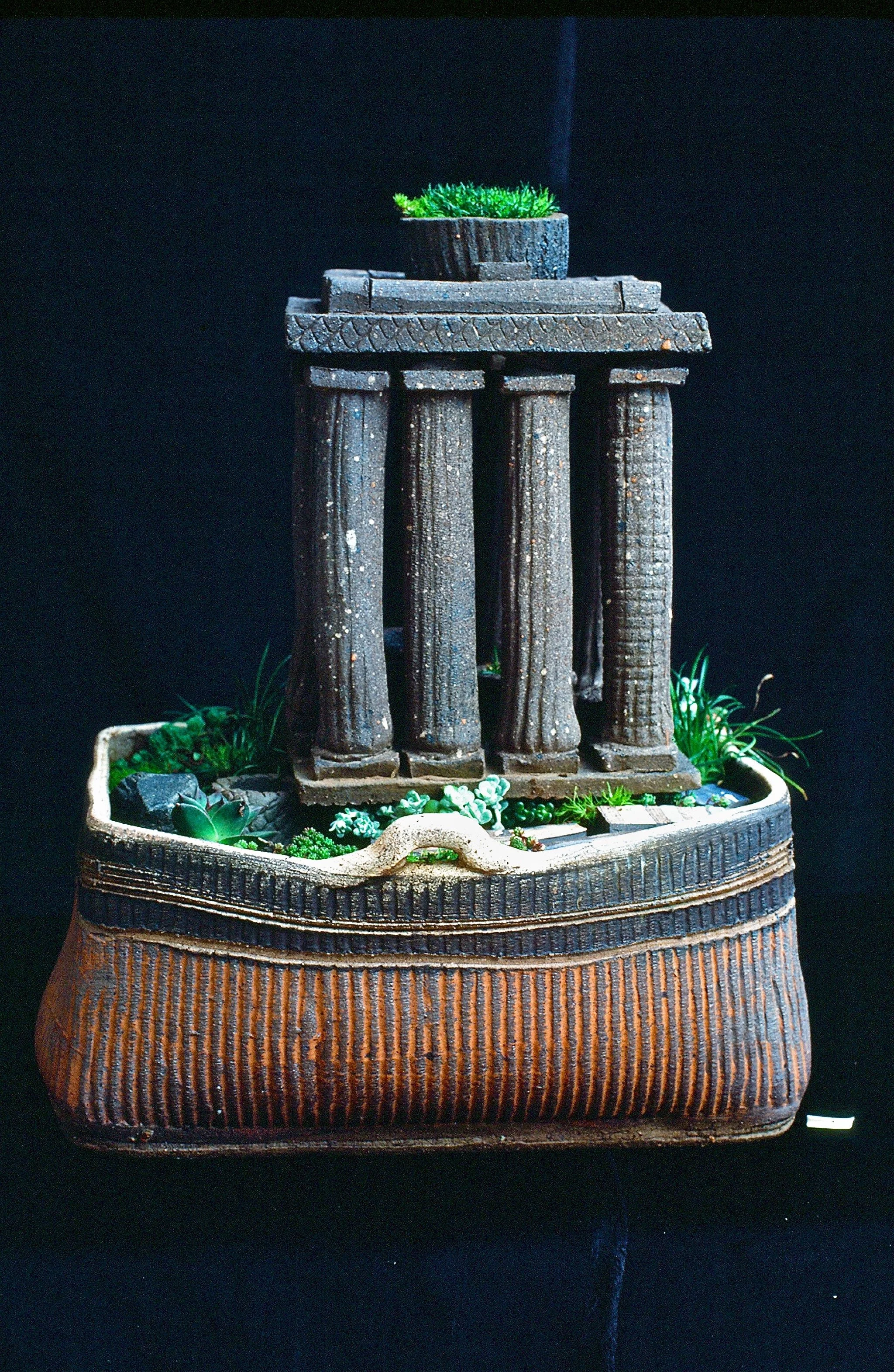 Small decorative fountain with columns, filled with artificial plants, set against a dark background.