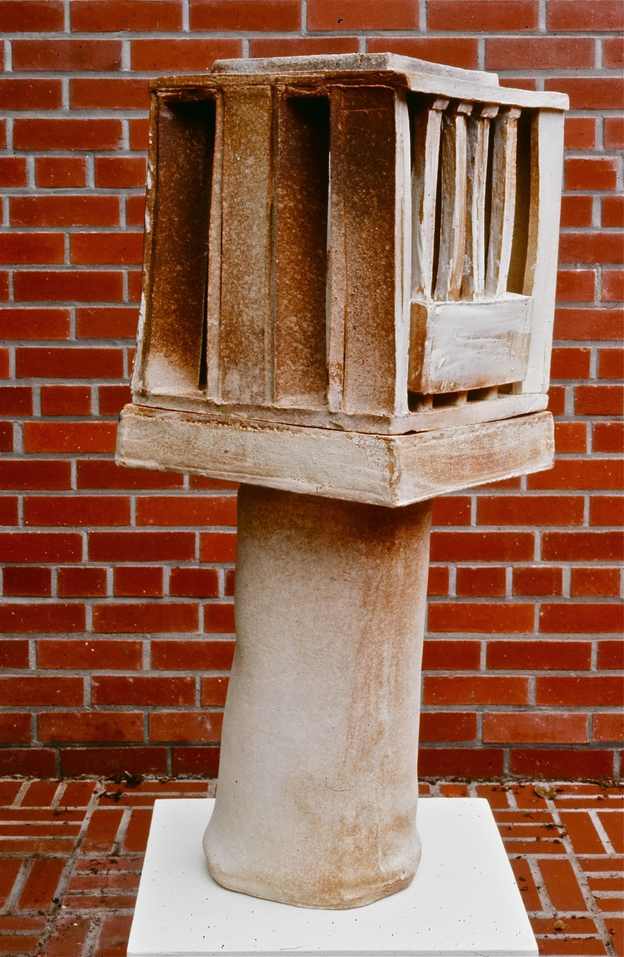 Ancient terracotta or stone artifact resembling a small temple or shrine with columns and a roof, mounted on a rounded pedestal, with a brick wall in the background.