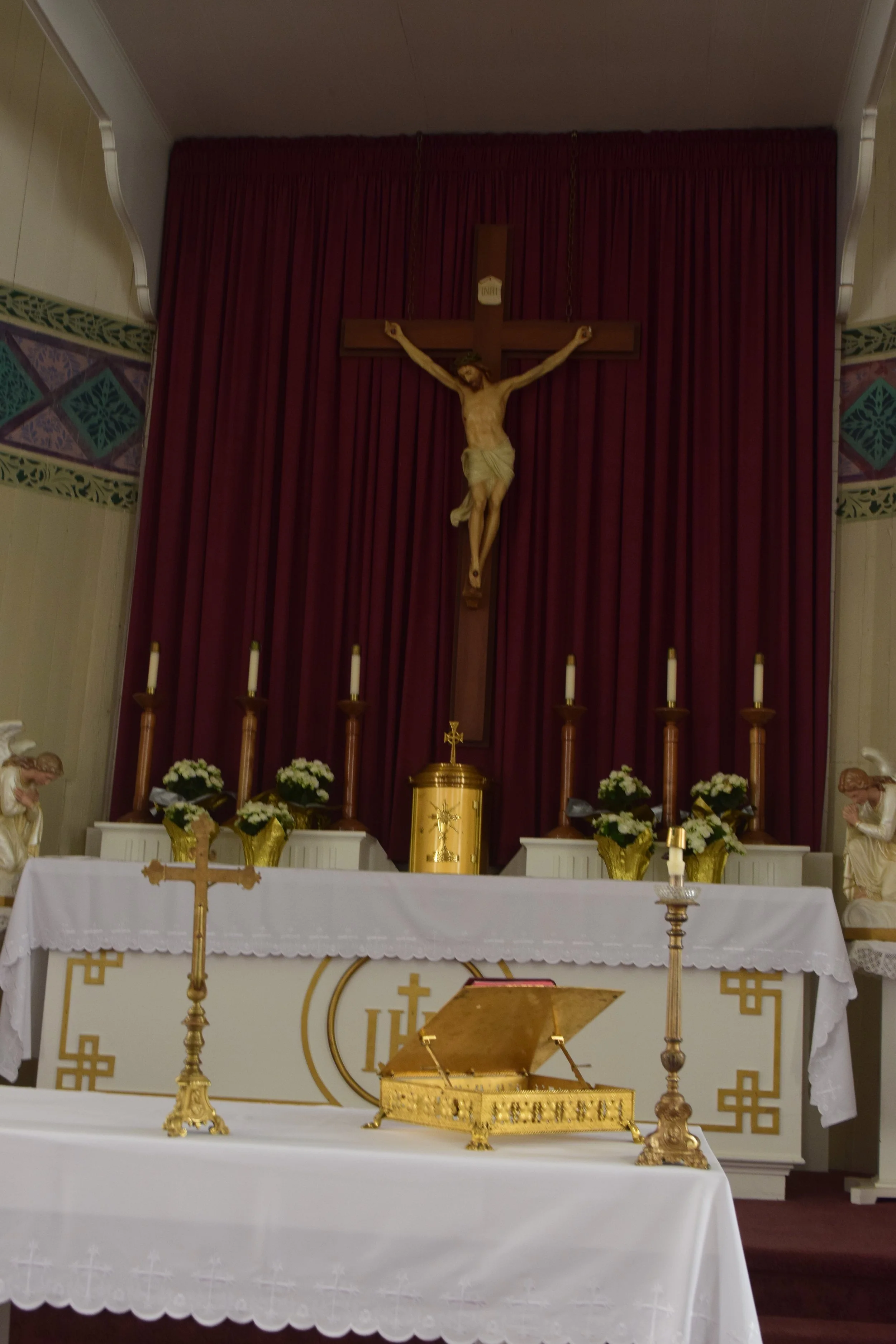 A church altar with a crucifix of Jesus Christ hanging on a wooden cross behind red curtains. The altar has white cloth with gold accents, and is decorated with white flowers in gold vases. There are five white candles placed on tall candle holders, 