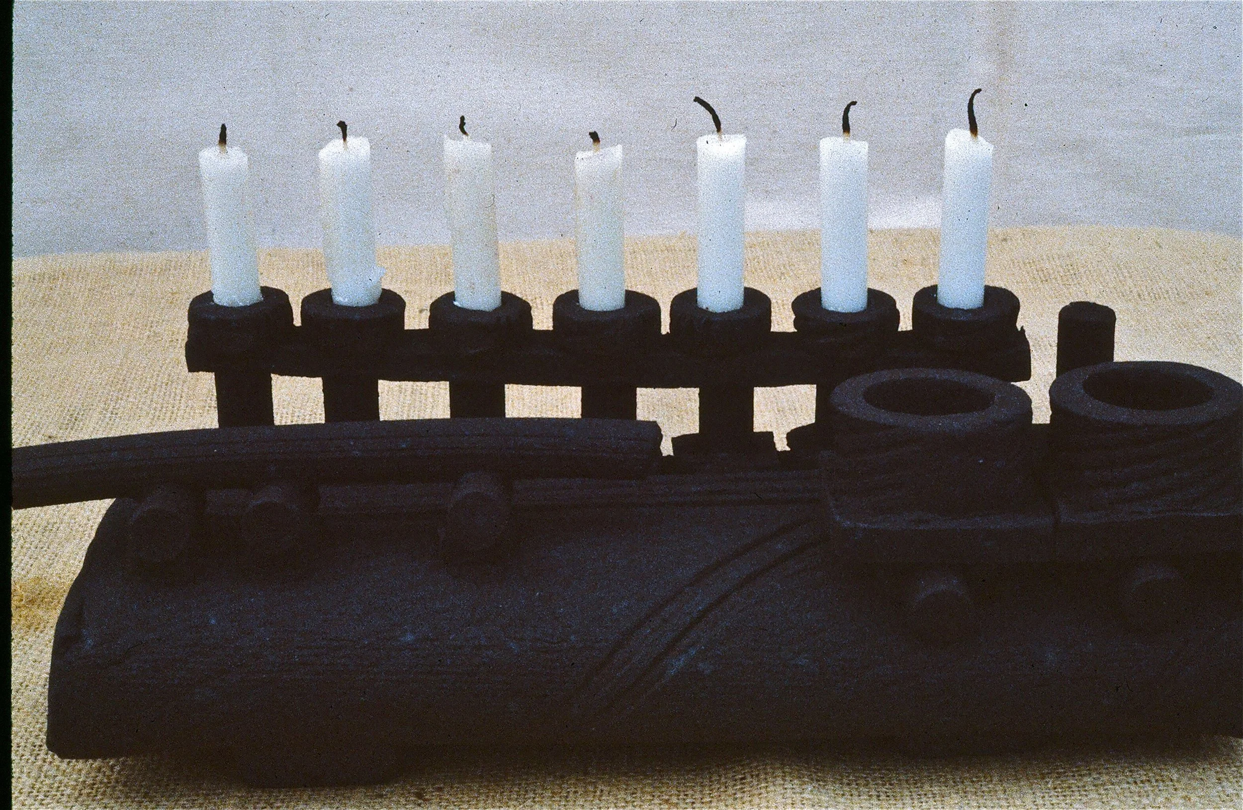A menorah with seven unlit white candles on a black stand, placed on a table with a beige cloth.