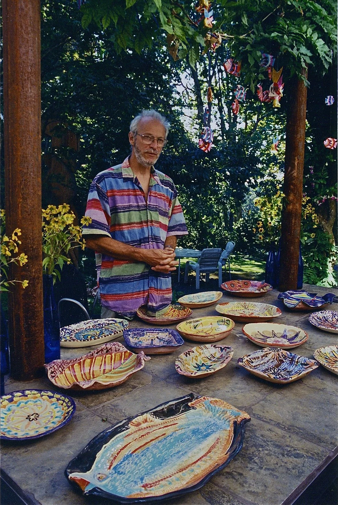 An older man with glasses and gray hair, wearing a colorful striped shirt, stands behind a table with various decorated ceramic dishes outdoors. The background features lush green trees and plants, with hanging colorful paper butterflies and flowers.