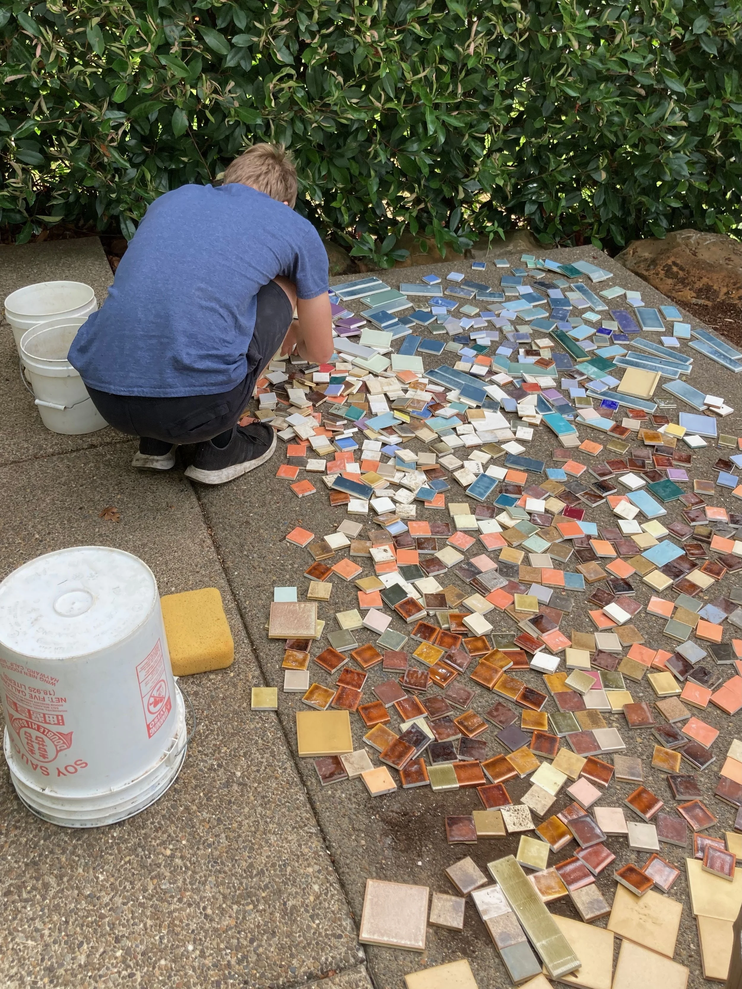A person crouching beside a large spread of colorful ceramic tiles laid on the ground, with three white buckets nearby and green bushes in the background.