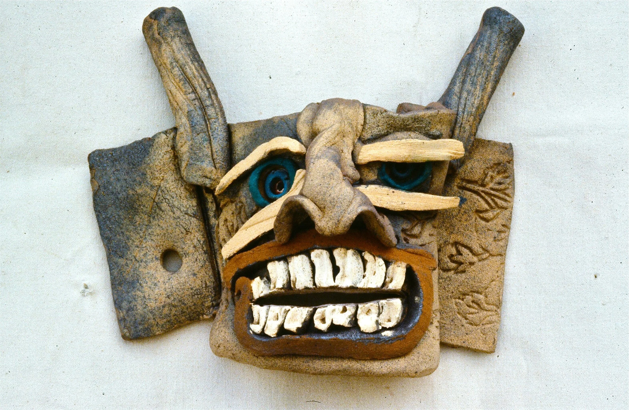 A colorful tribal mask made of various materials including wood and clay with exaggerated facial features, large ears, intense blue eyes, a prominent nose, and a wide open mouth showing teeth, set against a plain background.