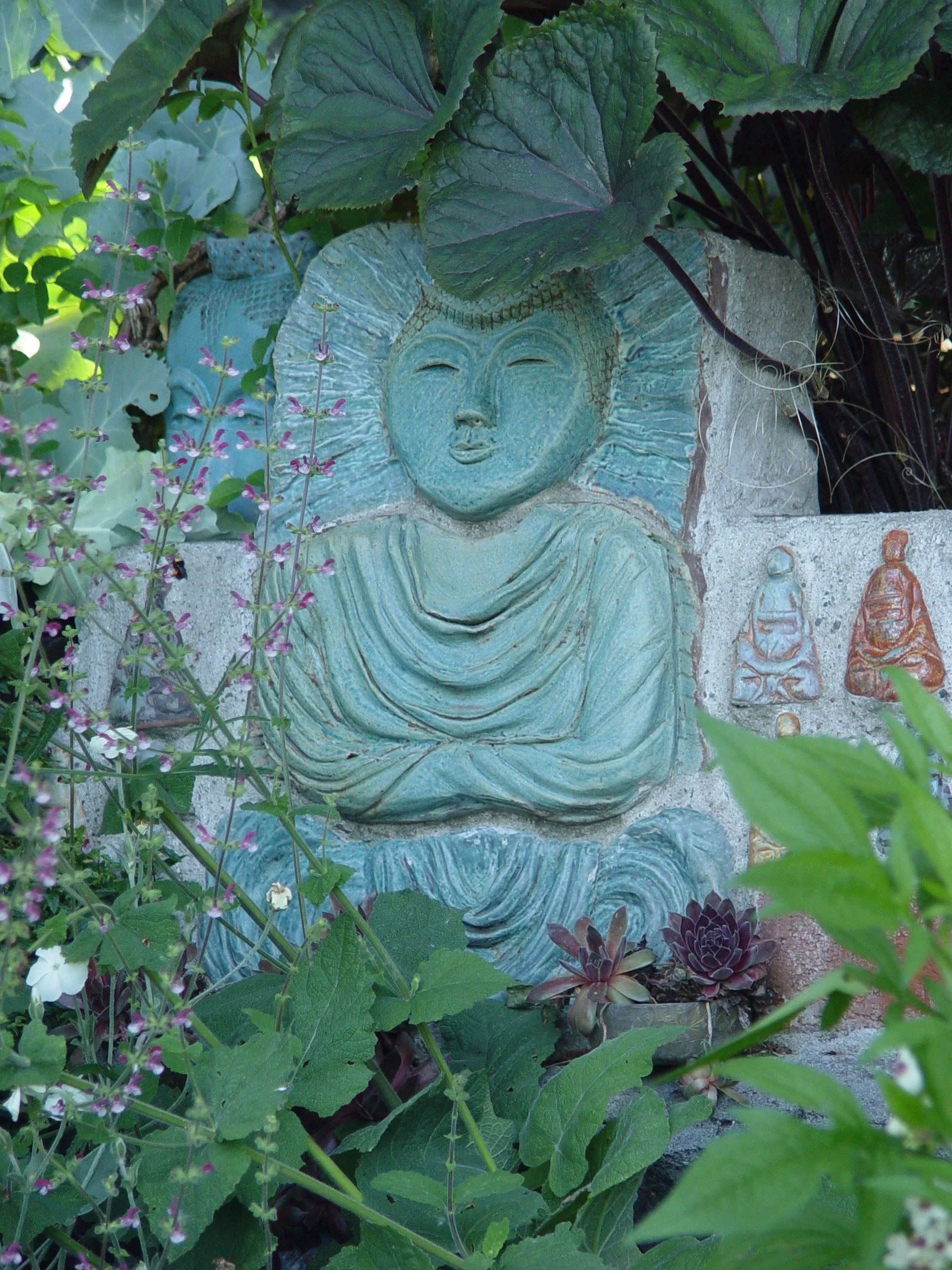 A carved stone or concrete Buddha statue with a serene facial expression, surrounded by green plants and purple flowers, in an outdoor garden setting.