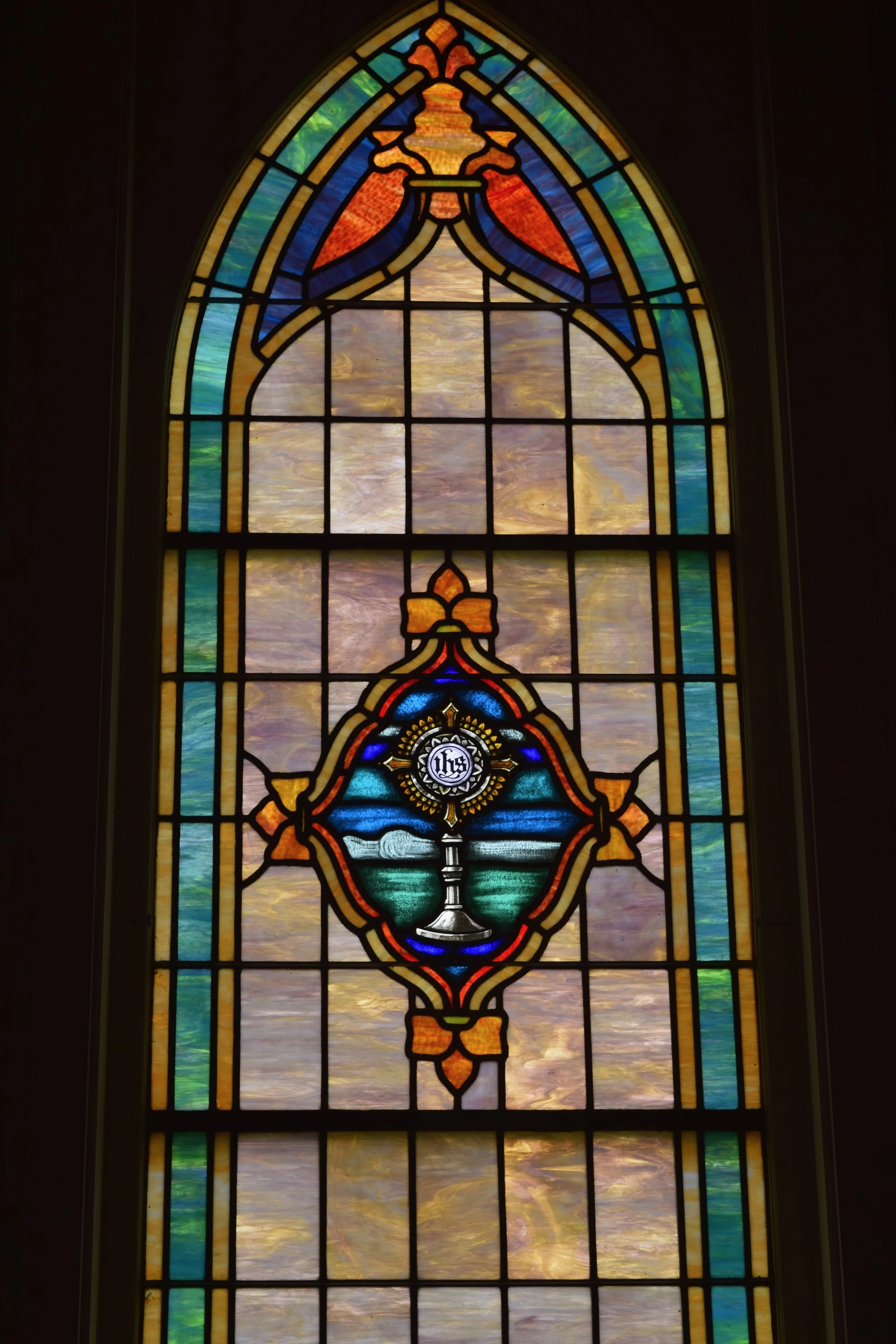Colorful stained glass window with a central emblem of a chalice, host with host initials, and decorative borders.