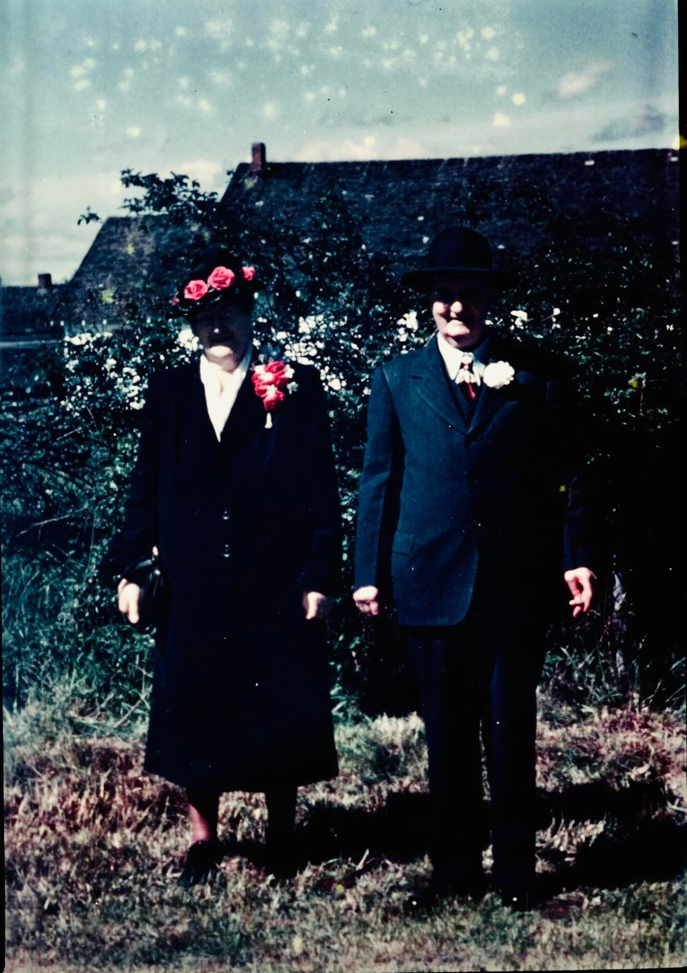 A black-and-white photograph of an elderly woman and man standing outdoors in front of bushes. The woman wears a dark coat and a hat decorated with pink flowers, while the man wears a dark suit with a white shirt, tie, and a bowler hat. Both have flo
