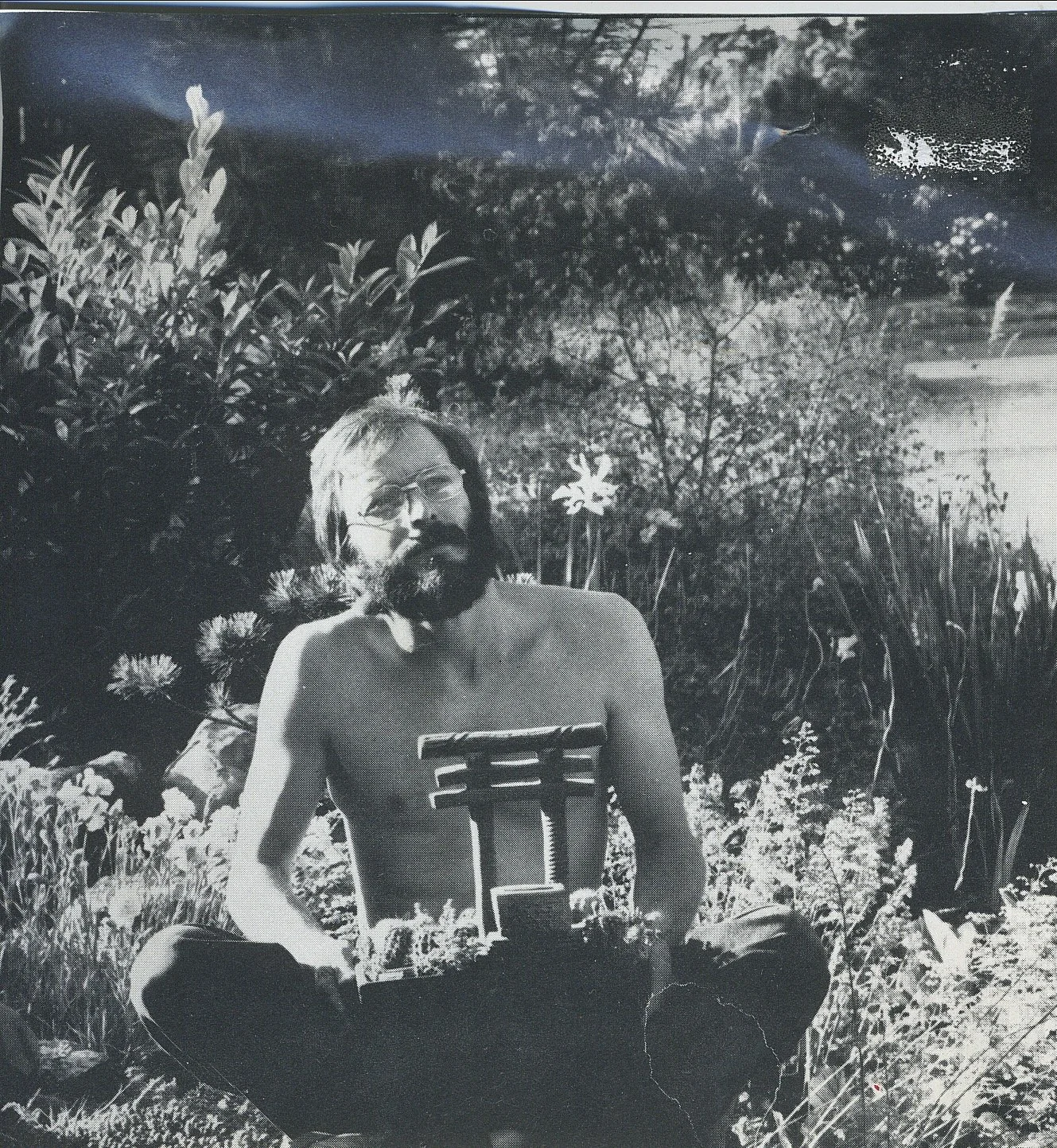 A shirtless man with glasses and a beard sitting outdoors among plants and flowers, with trees and sky in the background.