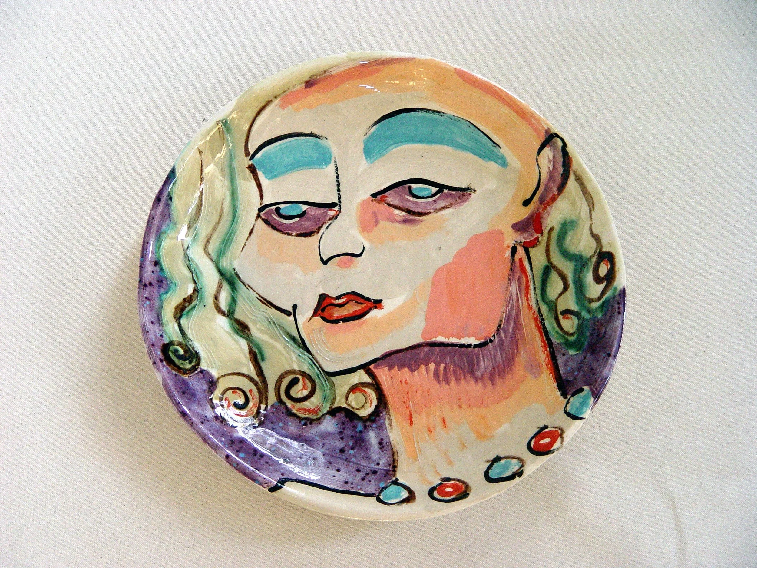 Decorative ceramic plate with abstract portrait of a woman with long wavy hair, blue eyeshadow, and red lips, featuring swirling patterns and pastel colors.