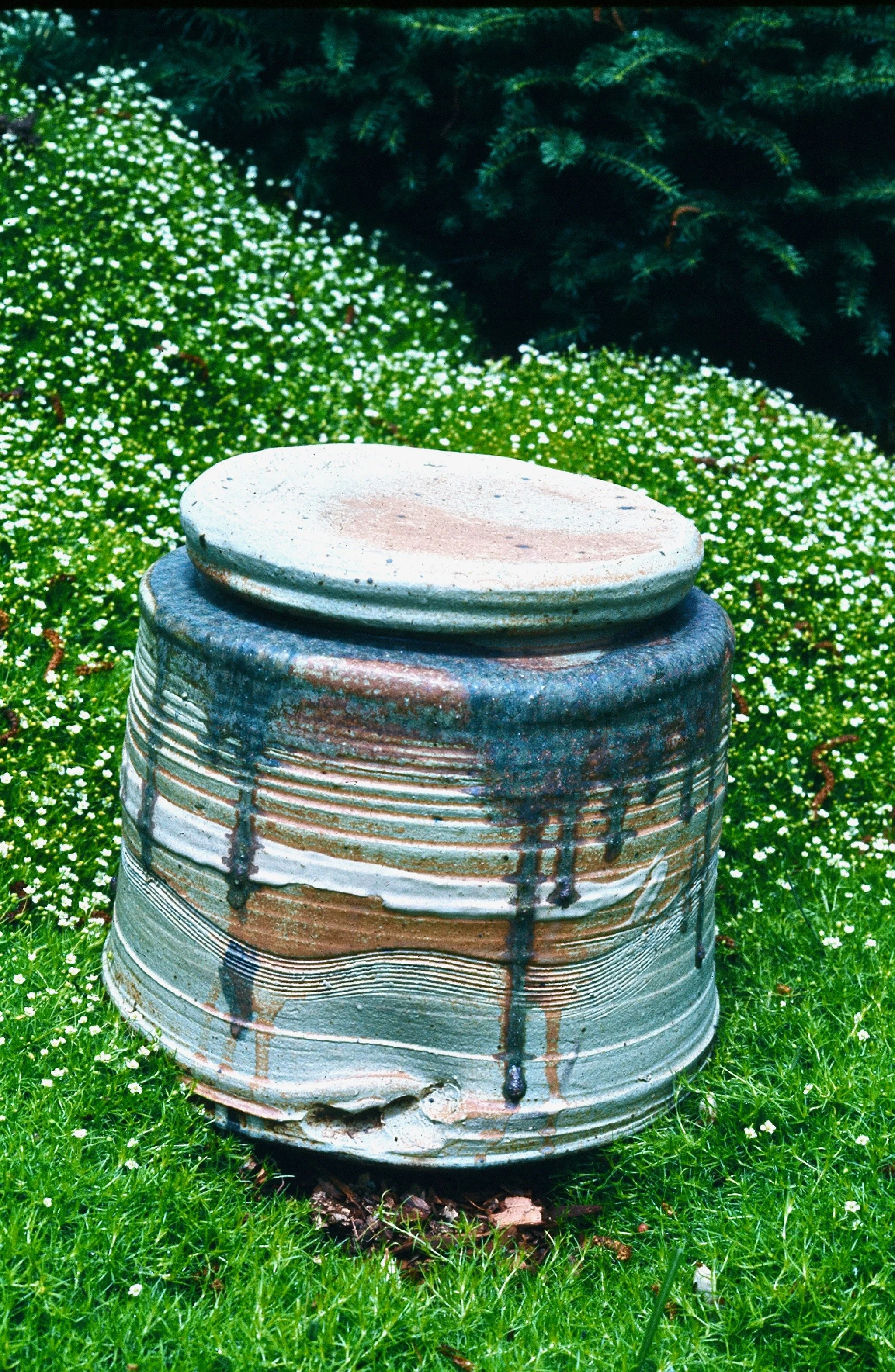 A large ceramic jar with a small flat dish on top sits outdoors on green grass surrounded by small white flowers, with trees in the background.