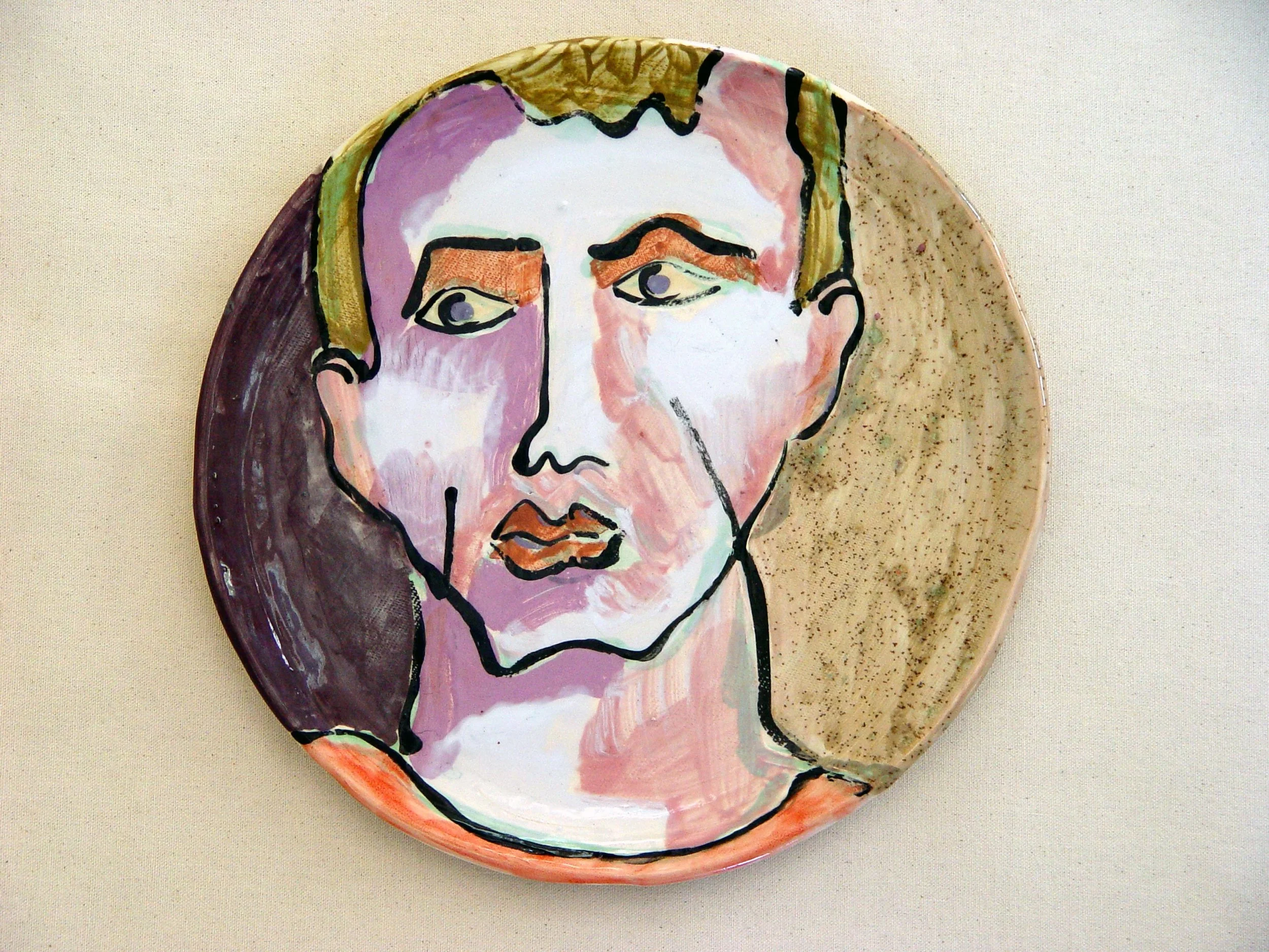 A decorative ceramic plate featuring a stylized abstract portrait of a person's face with bold black outlines, painted in shades of pink, purple, and beige, against a speckled beige background.