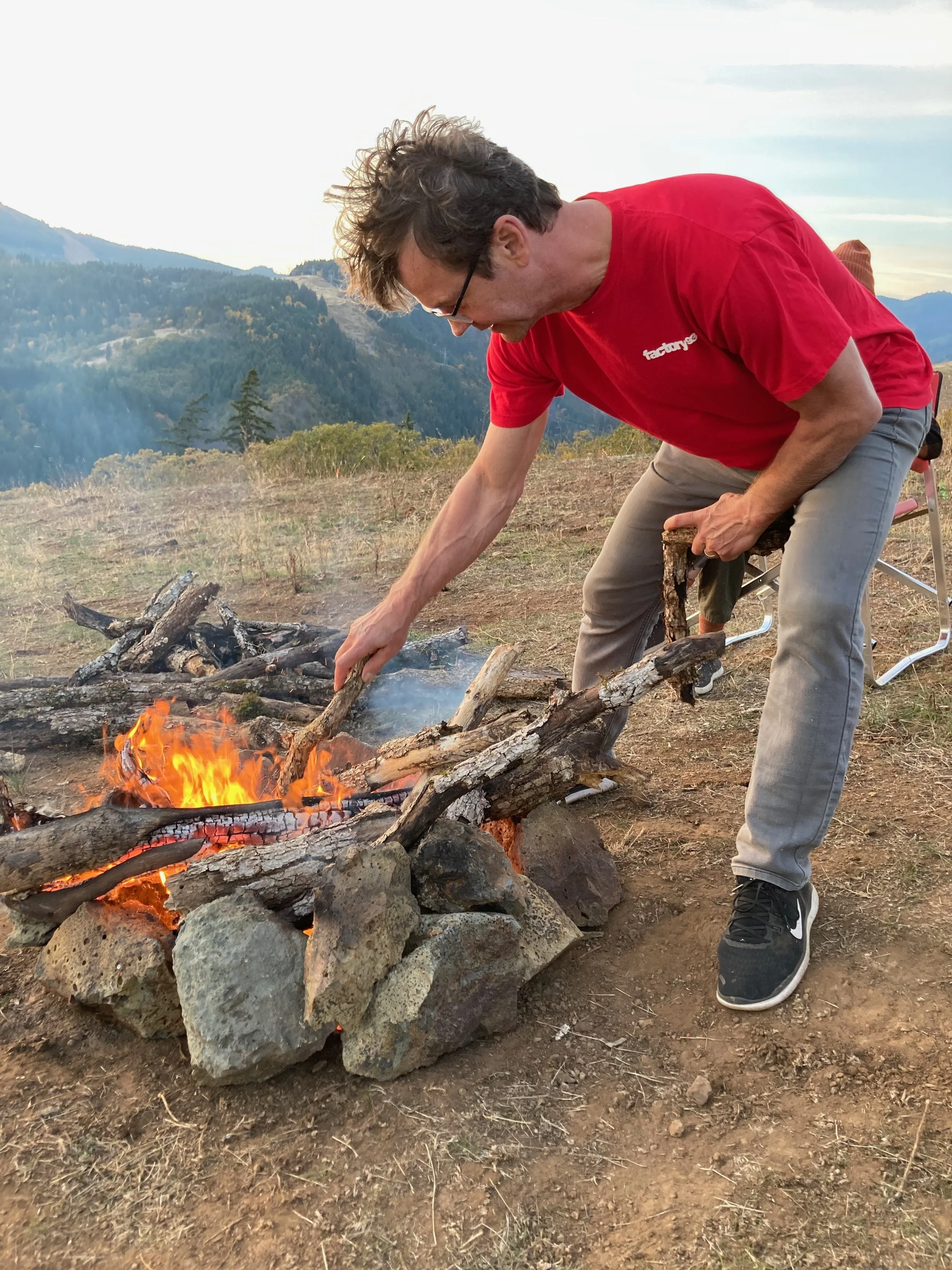 A man in a red shirt and gray pants arranging logs in a campfire outdoors on a mountain landscape during sunset.