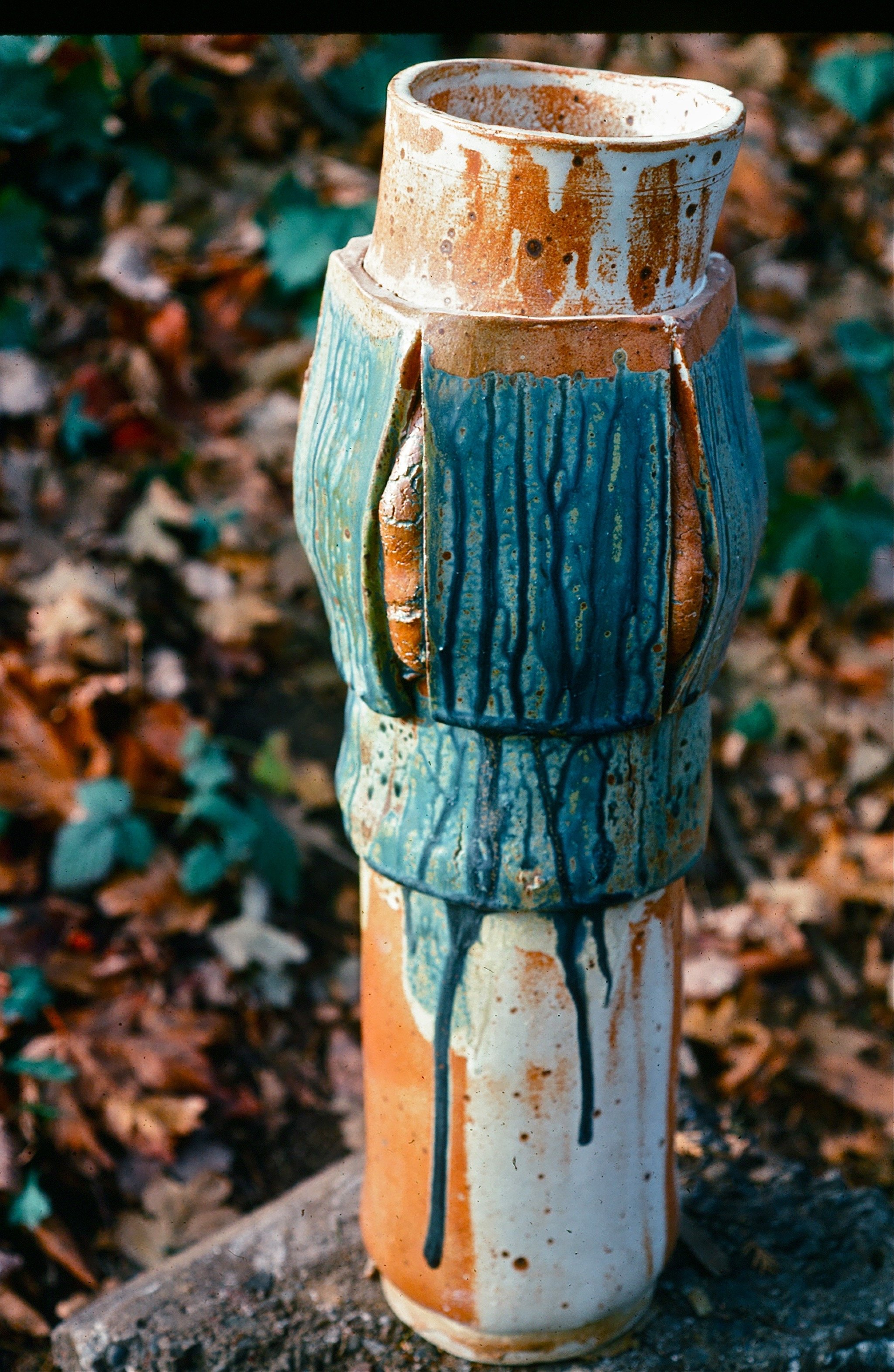 A ceramic sculpture of a human figure with a pipe-shaped head, painted in blue, white, and rust colors, outdoors on a dirt surface with fallen leaves and plants in the background.
