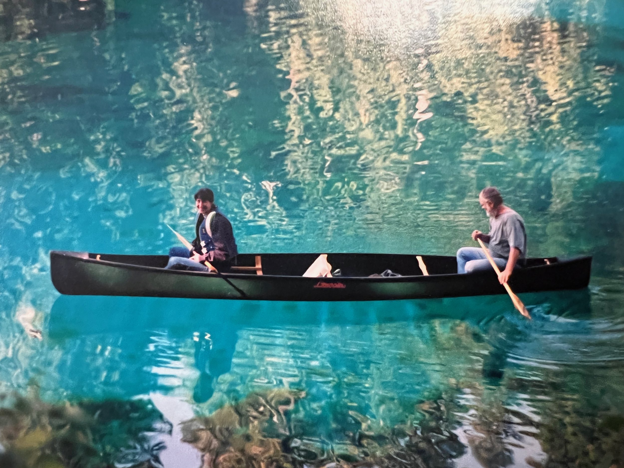 Two people in a black canoe on a calm, greenish-blue river, surrounded by trees.